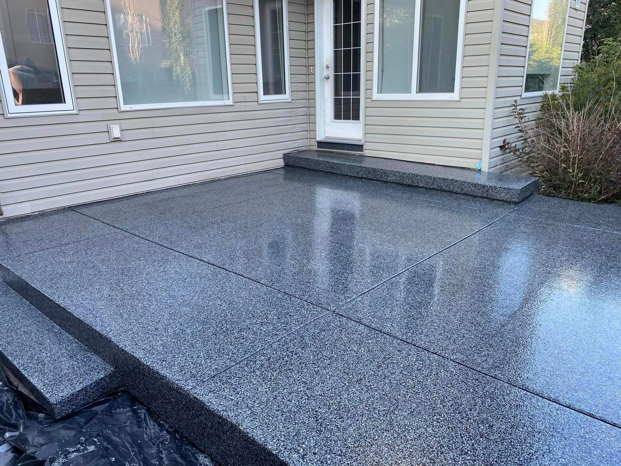 A newly finished gray speckled epoxy concrete patio outside a house with beige vinyl siding, large windows, and a front door, with some bushes on the right side.