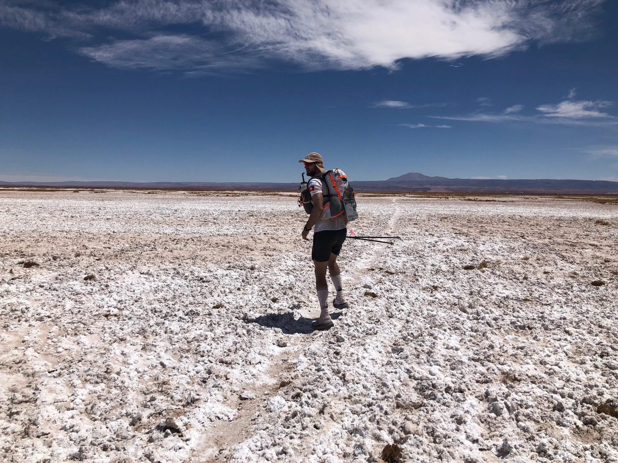 A person wearing a hat, sunglasses, shorts, and a backpack walking and running on a salt flat with a distant mountain and a partly cloudy sky.