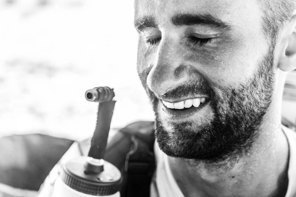 A close-up black-and-white photo of a smiling man with wet hair and a beard, holding a bottle with a homemade pipe or bong in a relaxed setting.