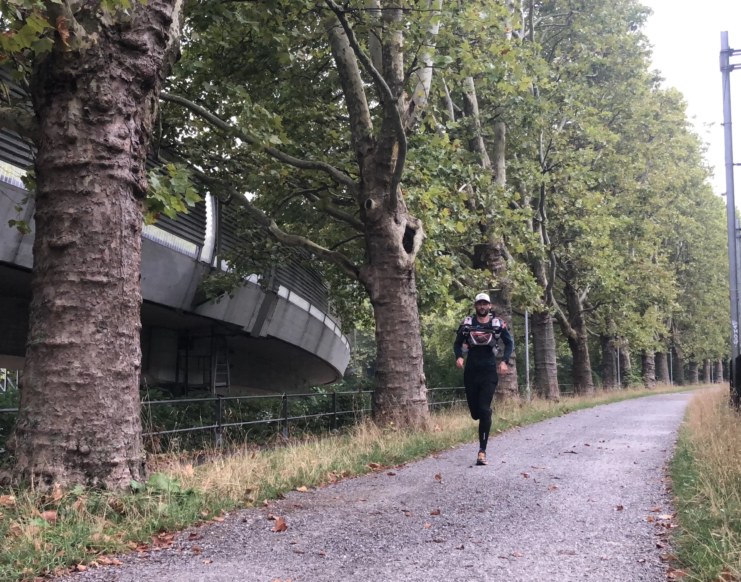 A man running along a gravel path lined with trees in an urban park. The trees have green leaves, and there is a metal fence along the side of the path. A streetlamp is visible on the right side of the image.