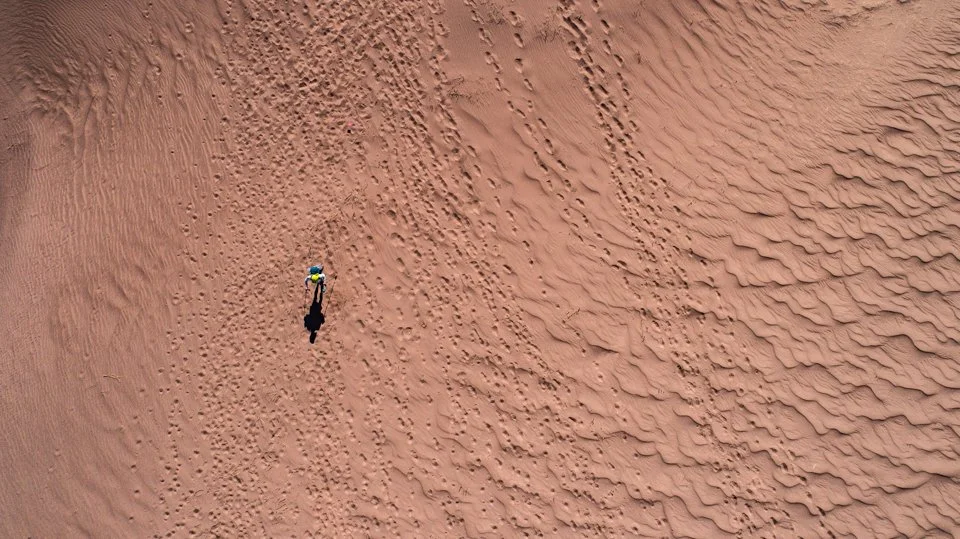 A person walking across a vast sandy desert with ripples and footprints.