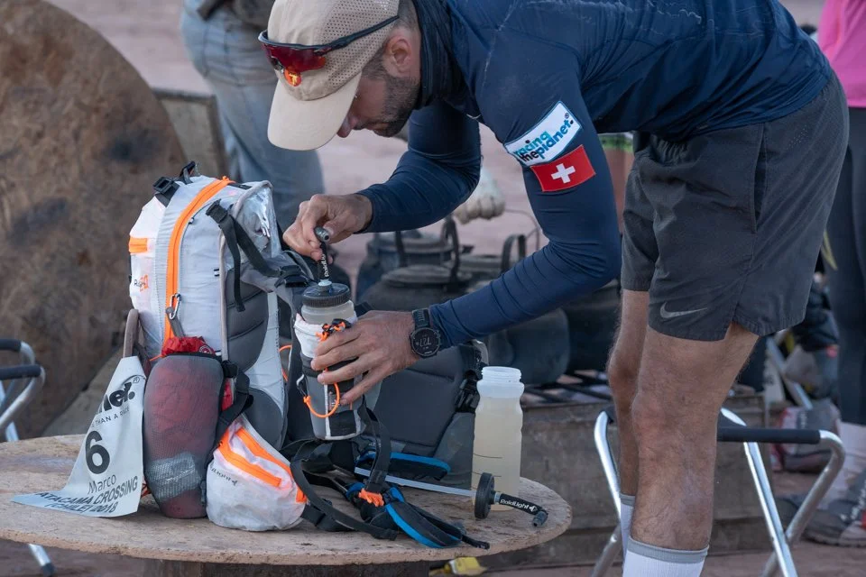 A man wearing a beige cap, sunglasses, a blue long-sleeve shirt with a Swiss flag patch, and shorts is filling a water bottle from a hydration pack. He appears to be preparing for a trail run or hike, with backpacks and gear on a wooden table.