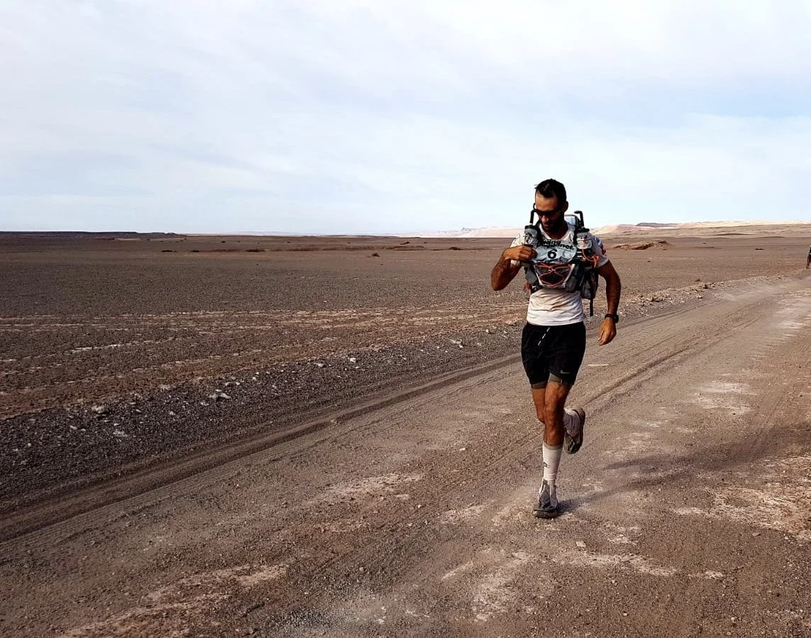 A man running on a dirt road in a desert landscape, wearing athletic clothing and a hydration pack.