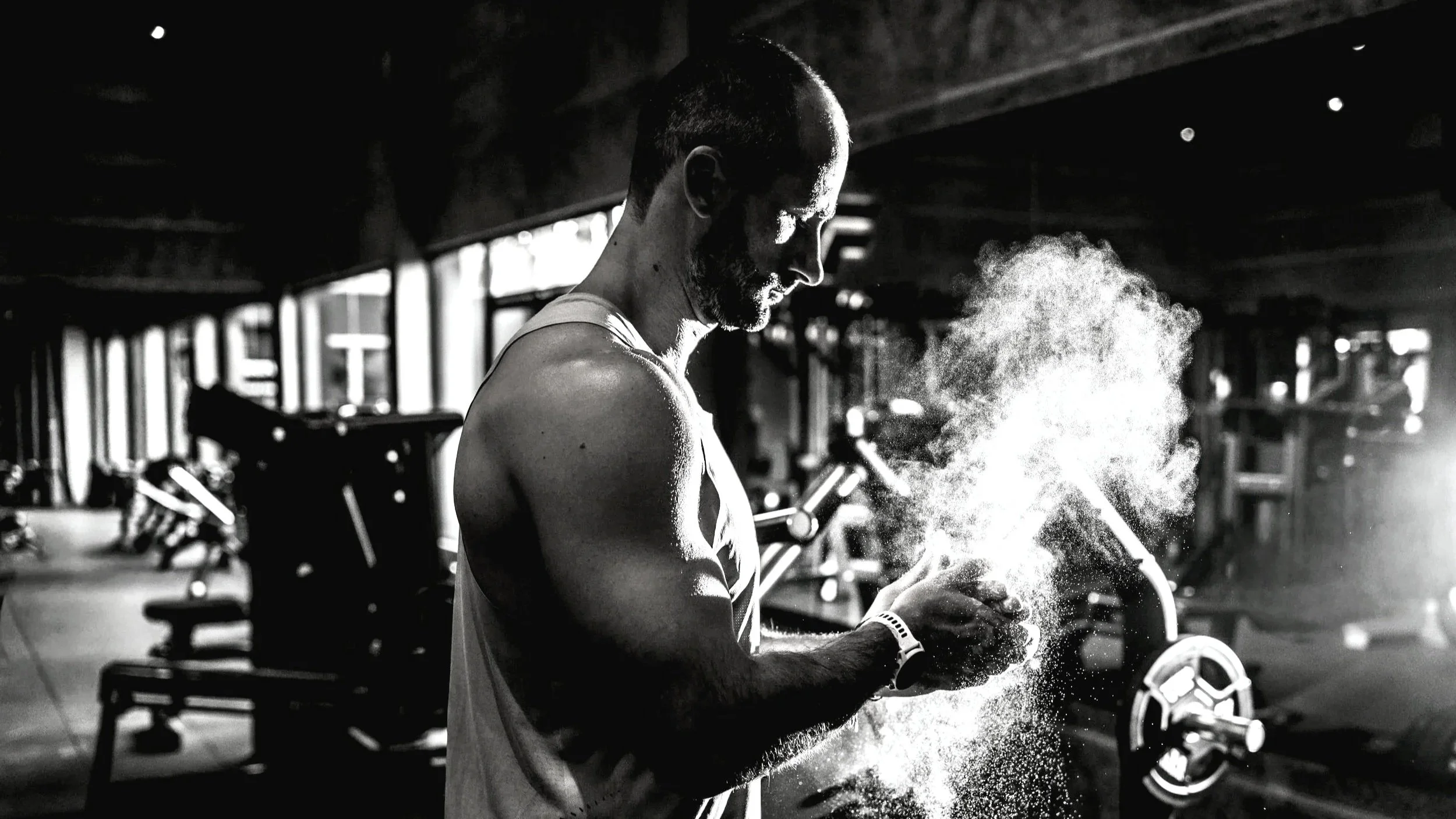 A man in a gym, cleaning his hands and surrounded by chalk dust with gym equipment in the background.