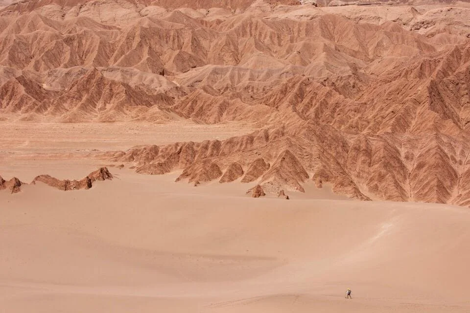 Desert landscape with reddish sandstone mountains and a person walking in the sand.