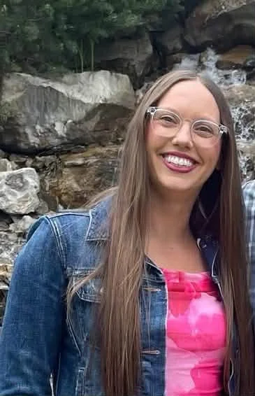 A woman with long brown hair and glasses smiling outdoors in front of rocks and a small waterfall.