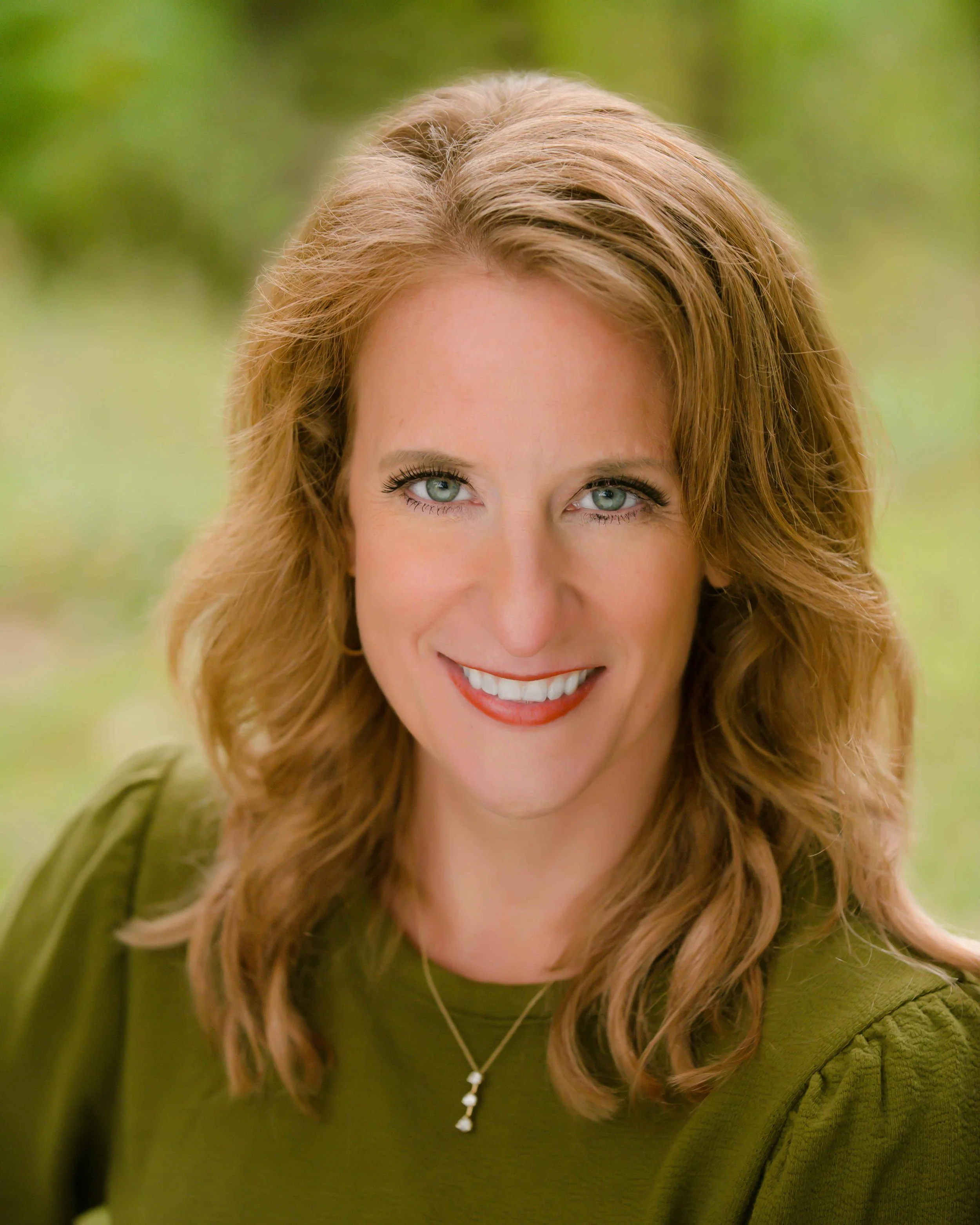 A woman with wavy, shoulder-length reddish hair and blue eyes, smiling, wearing a green top and a silver necklace with a small pendant, in an outdoor setting with a blurred green background.