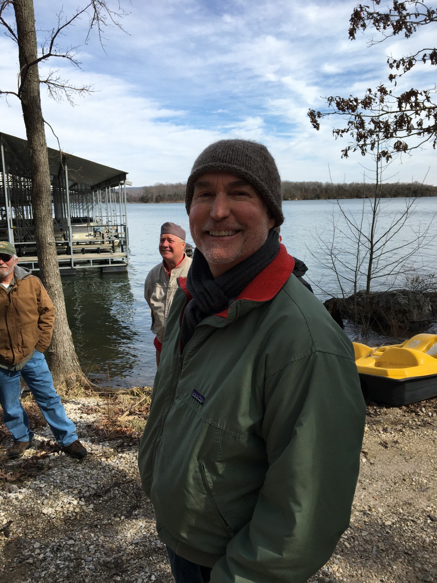 Man smiling outdoors near a lake, wearing a green jacket, gray beanie, and scarf, with two other men in the background and a yellow pedal boat on rocky ground.
