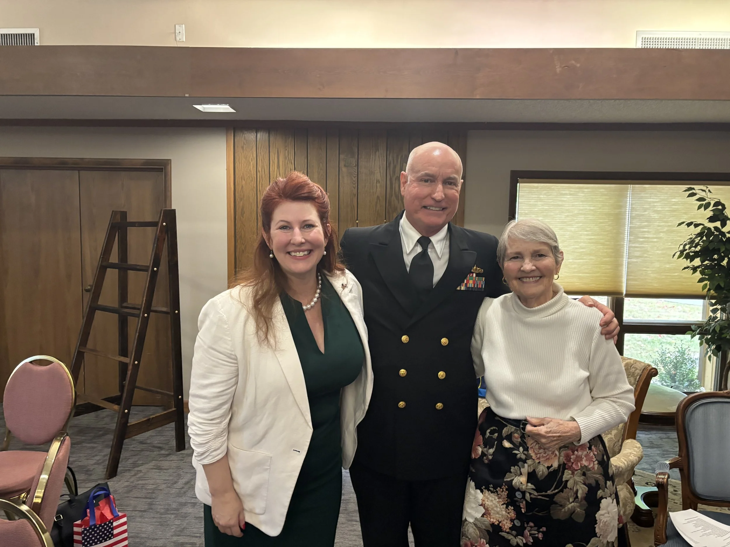 Three people standing together indoors, smiling at the camera. The person in the middle is a man in a military uniform. The woman on the left has red hair, wearing a green dress with a white blazer. The woman on the right has gray hair, wearing a white turtleneck and a floral skirt.