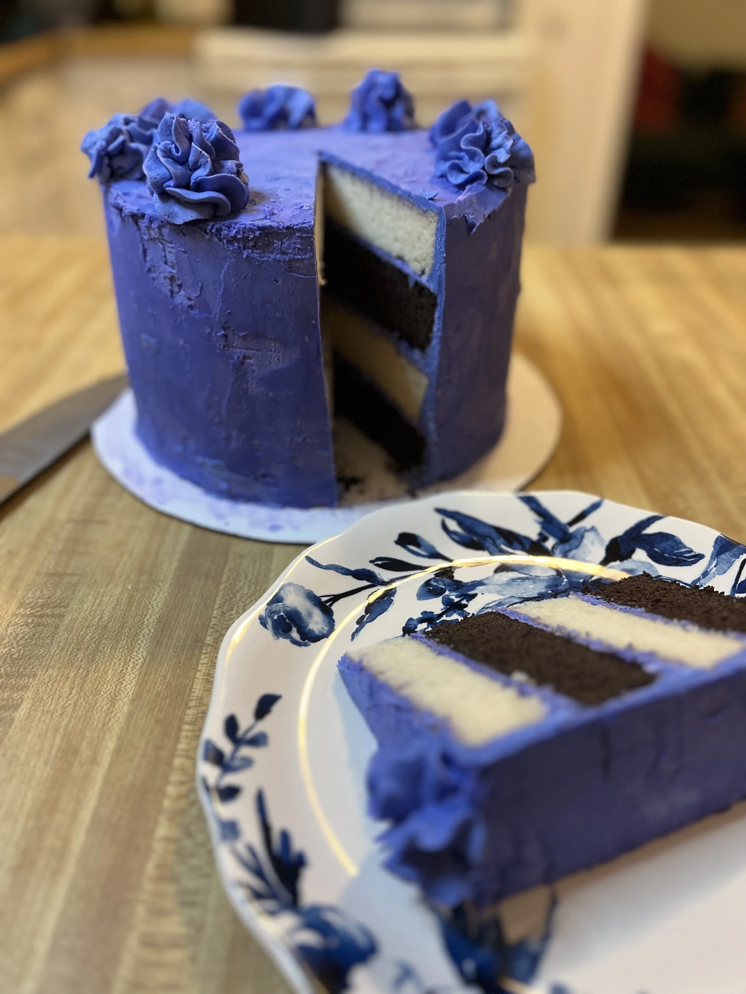 A partially sliced, three-layer, purple-frosted cake with decorative purple frosting accents on top, sitting on a white cake board on a wooden table, with a slice served on a matching blue-and-white floral plate.