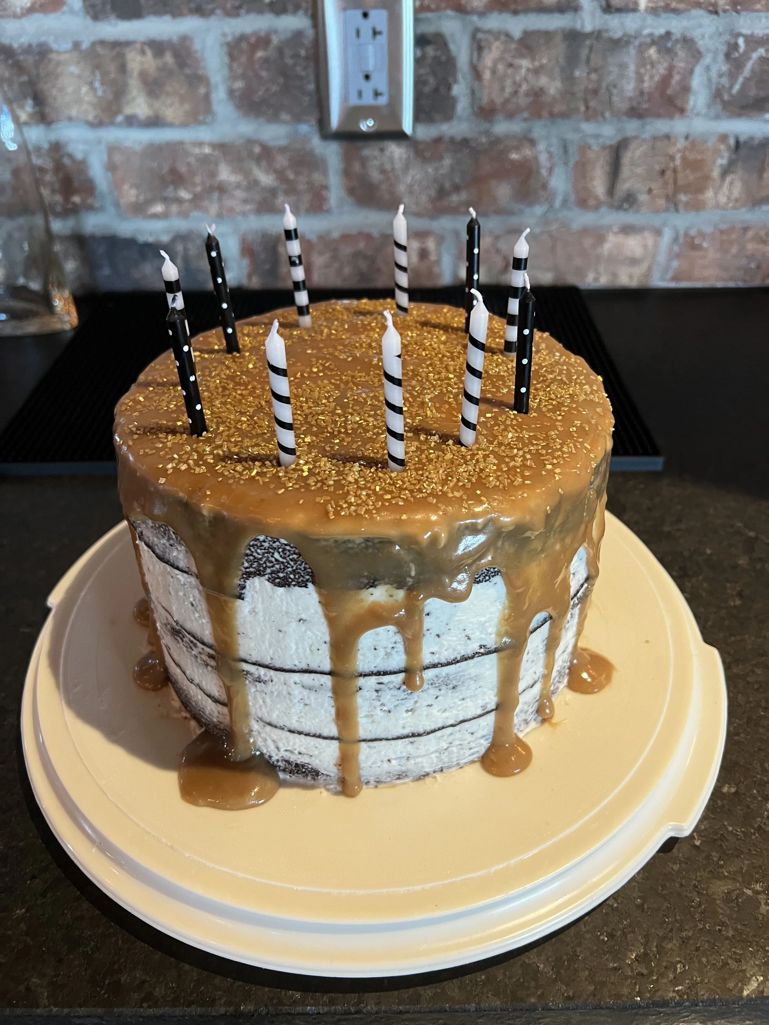 A layered birthday cake with caramel sauce dripping down the sides, topped with black and white striped candles, and gold sprinkles on top, placed on a white plate.
