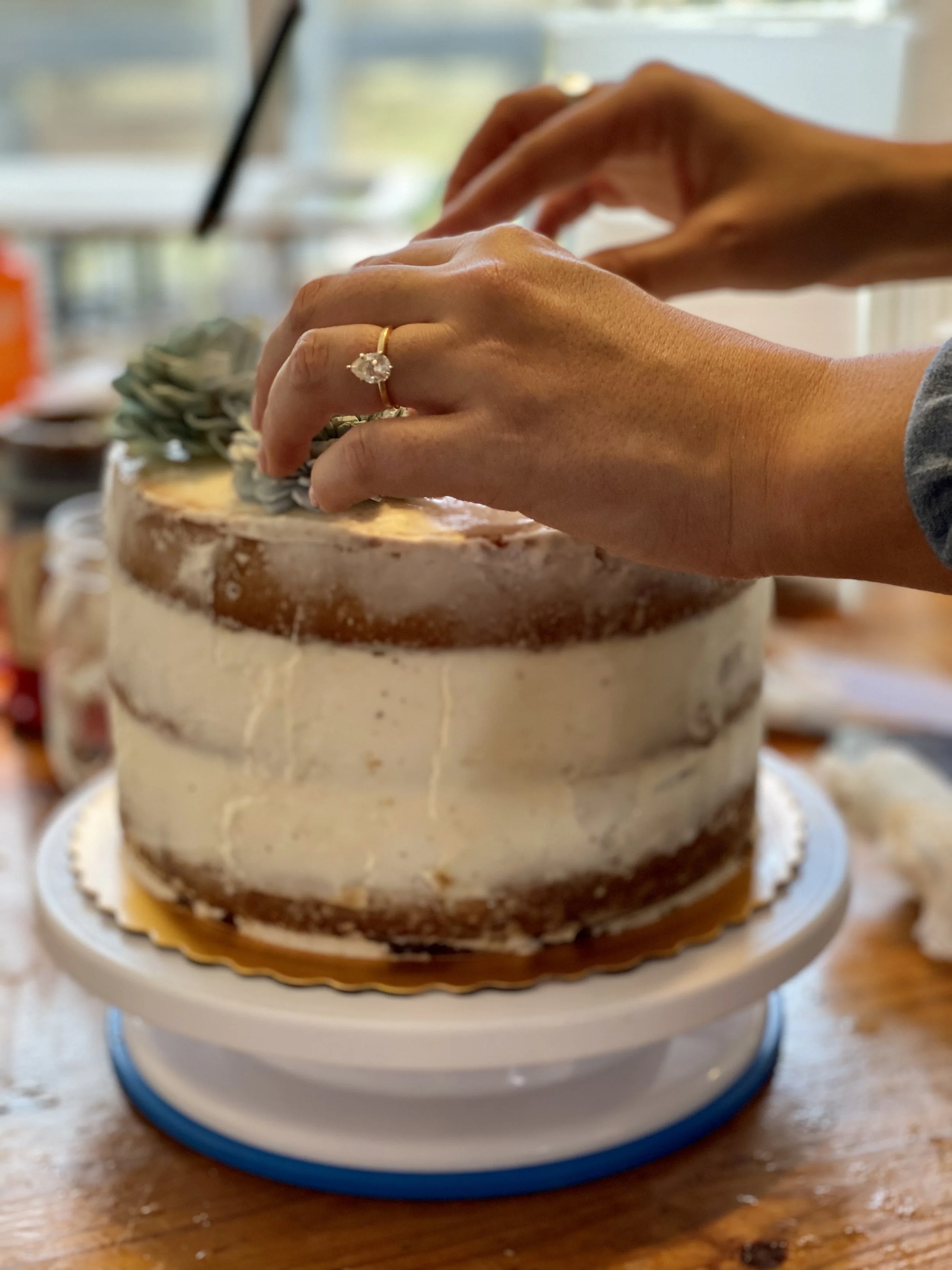 A person decorating a semi-naked cake with buttercream frosting, wearing a ring with a large gemstone. The cake is on a cake stand on a wooden table.