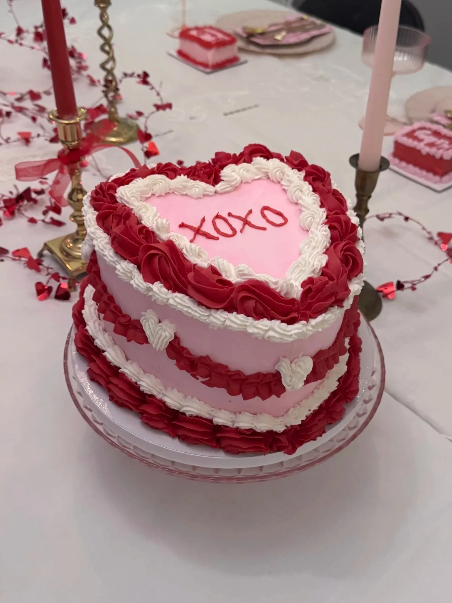 Heart-shaped cake with pink frosting, red and white decorative icing, and 'XOXO' written on top, on a dining table with pink candles and Valentines Day decorations.