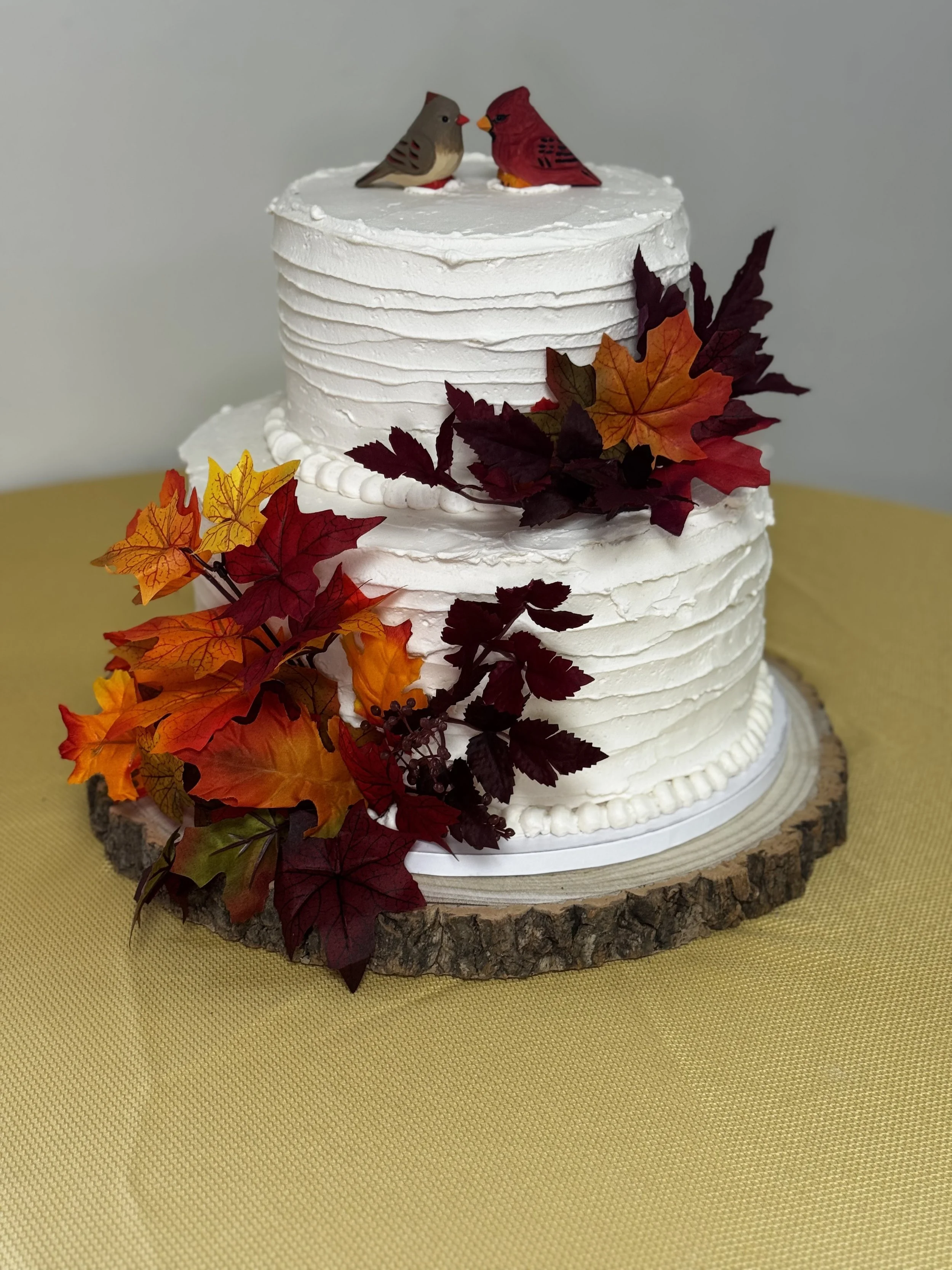 A two-tier white cake with leaf decorations and two bird figurines on top, placed on a wooden slab. Fall wedding cake.
