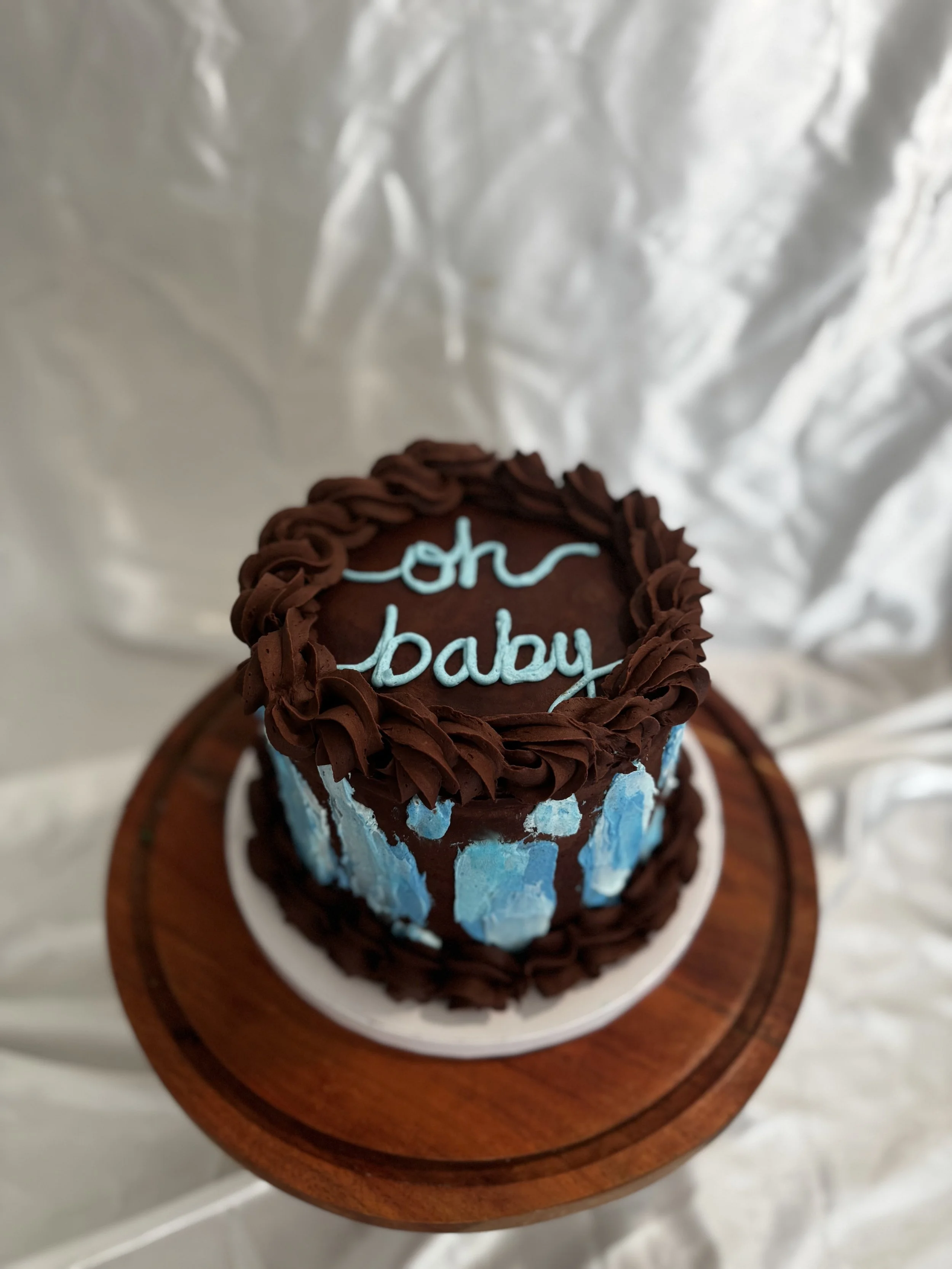 A round cake with blue and brown icing, decorated with a chocolate border and blue ice cream-like patches, has "oh baby" written in blue icing on top. It is placed on a white plate on a small wooden round stand. Baby Shower cake.