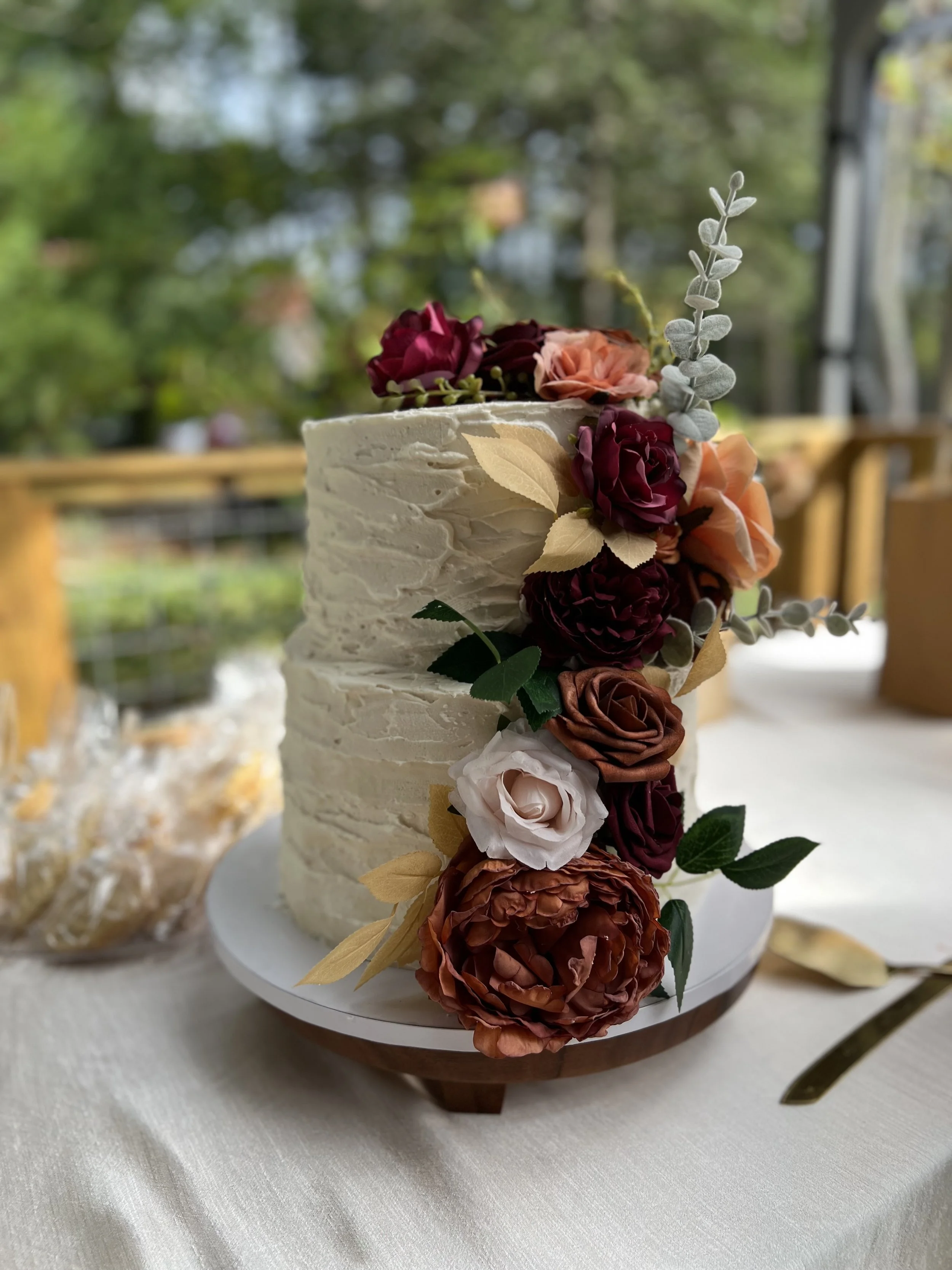 Two-tiered wedding cake with white textured frosting, decorated with pink, red, white, and brown flowers, and green leaves, on a white cake stand on a table with a white tablecloth.