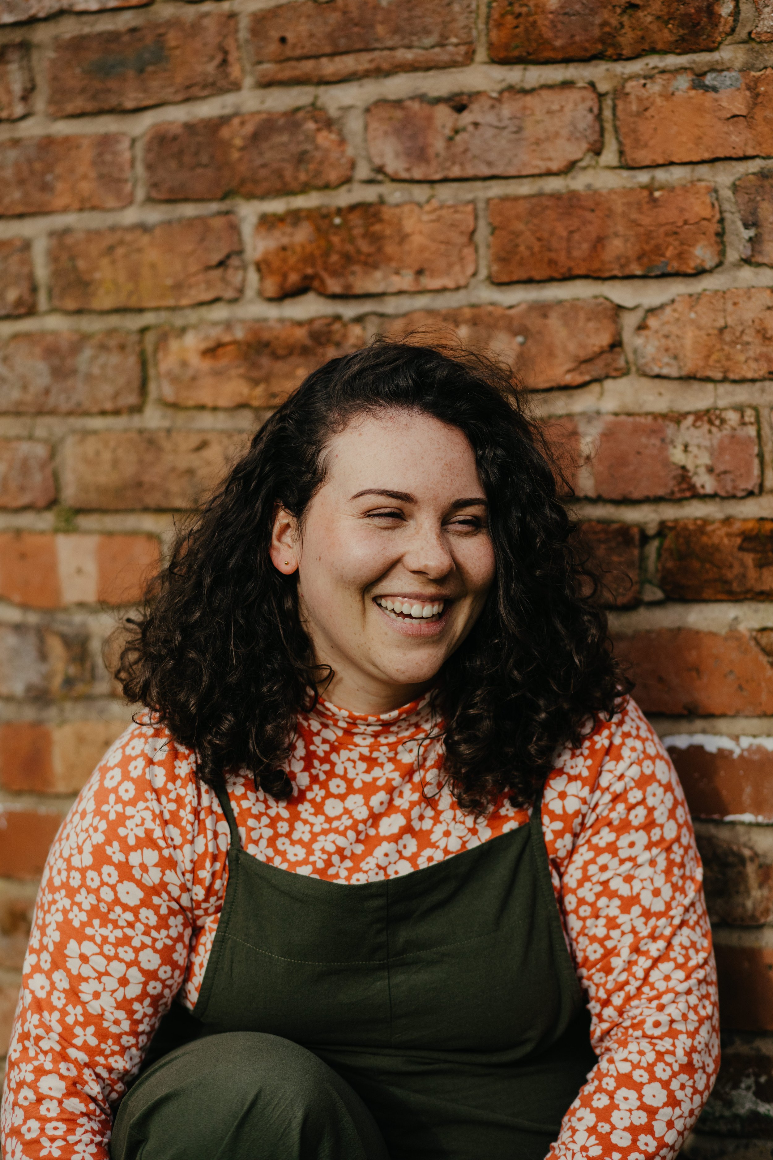 A woman with curly dark hair, smiling while sitting against a brick wall, wearing an orange and white floral top and a green apron.