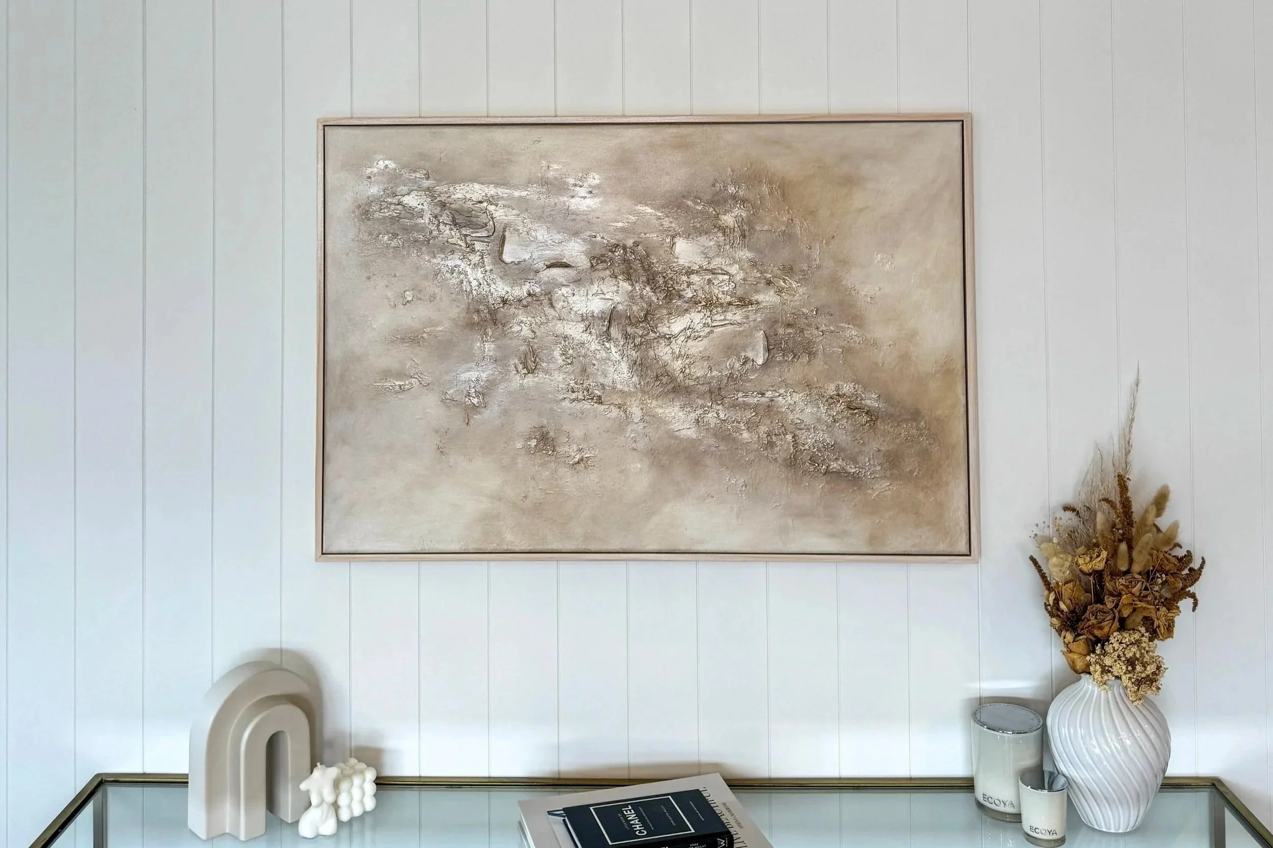 A framed abstract gold painting on a white wall, with a decorative white vase filled with dried flowers and grasses on the right side, and a white sculpture and a few books and candles on a glass table in front.