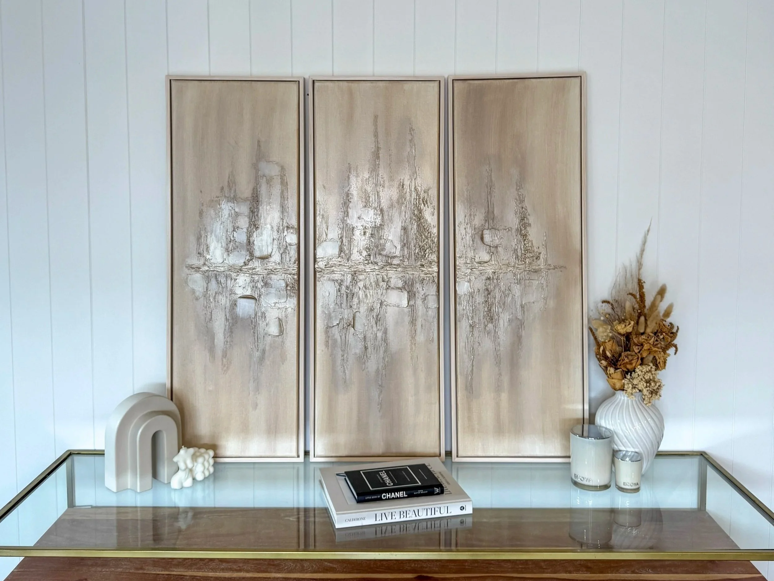 Decorative arrangement on a wooden table with books, candles, and a vase of dried flowers in front of a split panel gold art piece.