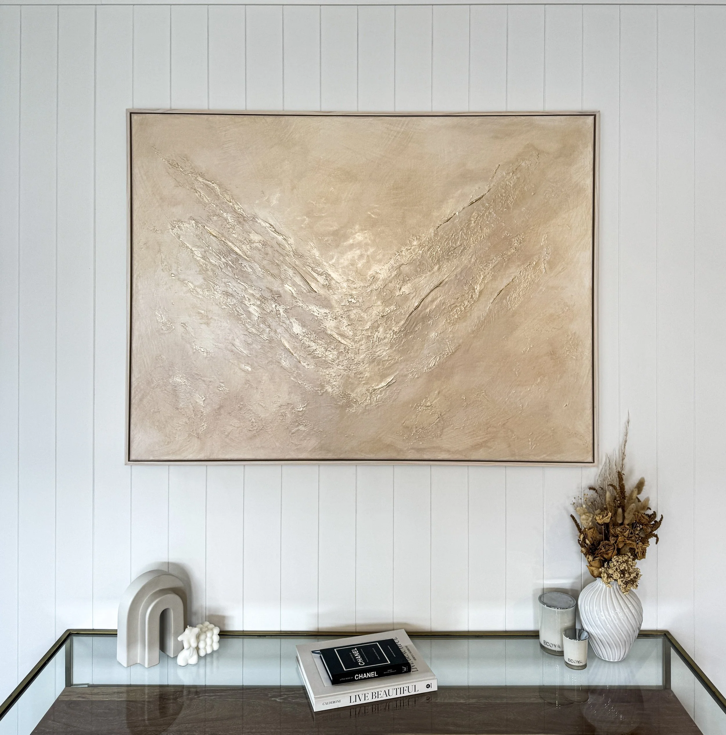 Decorative interior with a large abstract gold painting on a white panelled wall, a glass-top table with a white sculpture, a stack of books, and a white vase with dried flowers.
