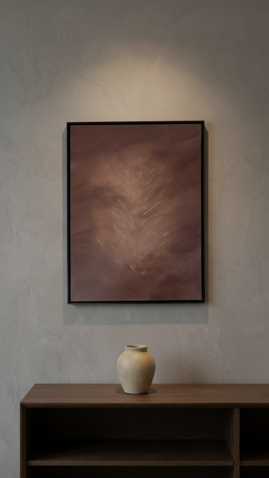 A beige vase on a wooden table with a framed abstract painting with a fern leaf design on the wall behind it