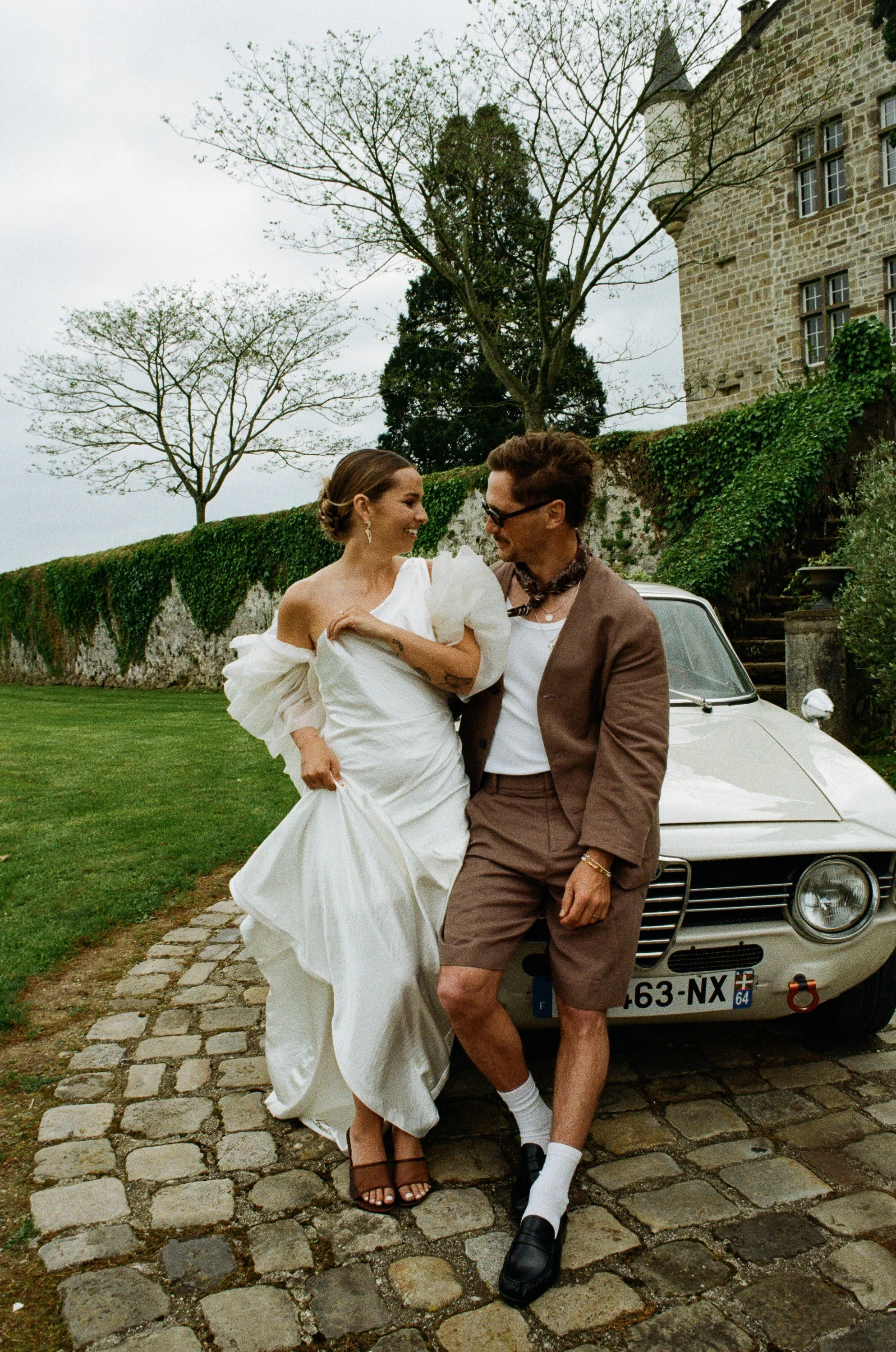 A couple stands in front of a classic white car, with the woman dressed in a wedding gown and the man in a casual suit, at a historic stone mansion with a stone path, green grass, and bare trees in the background. Taken on 35mm analogue film.