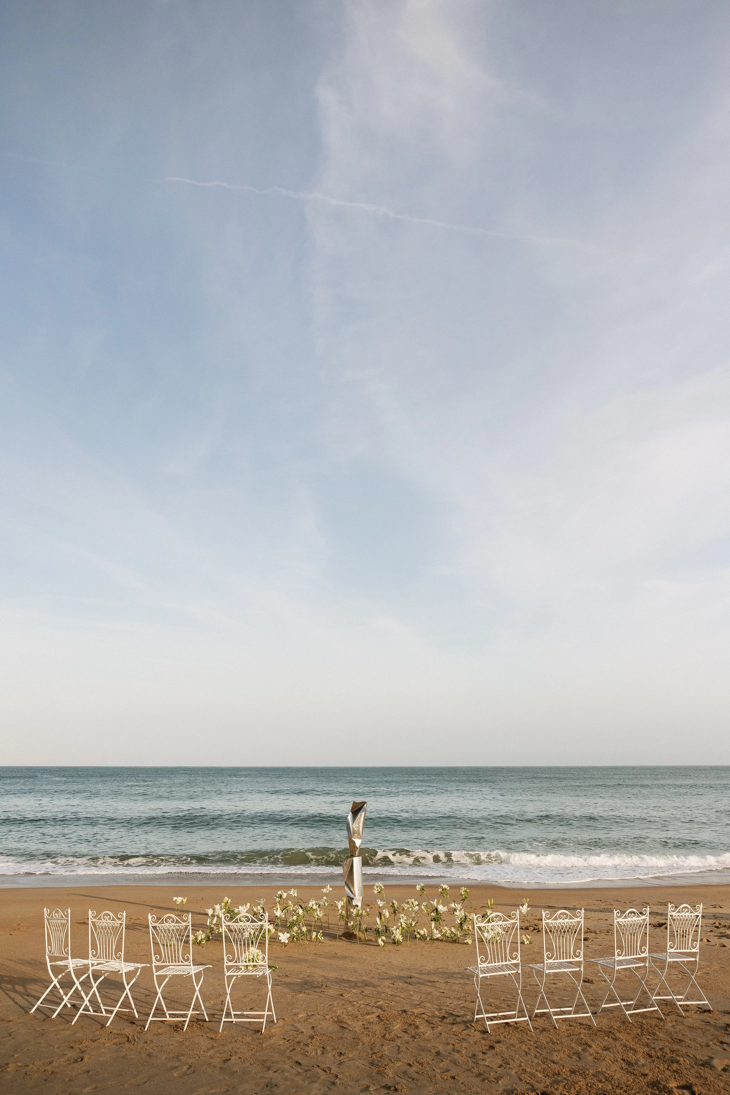 Beach scene with white chairs, flowers, and view of the ocean under a cloudy sky.