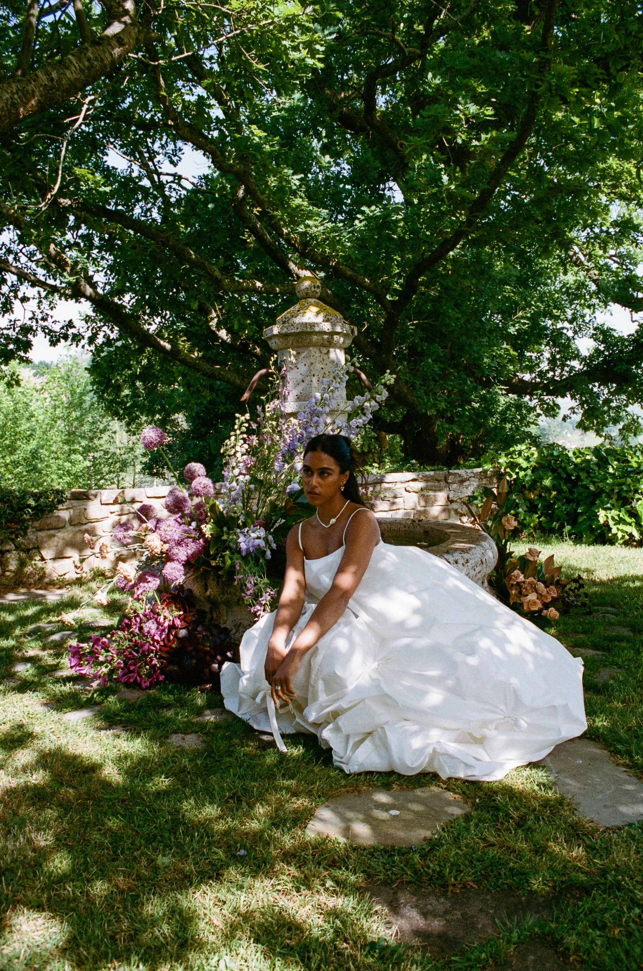 A woman in a white dress with spaghetti straps is sitting outdoors on a stone surface, surrounded by a large variety of colorful flowers including pink, purple, and white blooms. She is positioned near a stone structure and lush green trees provide shade in the background.