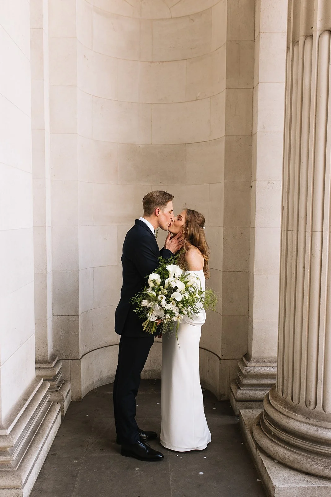 A man and woman in wedding attire sharing a kiss, standing in front of large stone columns. The woman holds a bouquet of white flowers, and they are in a classical architectural setting. The Old Marylebone Town Hall in London.
