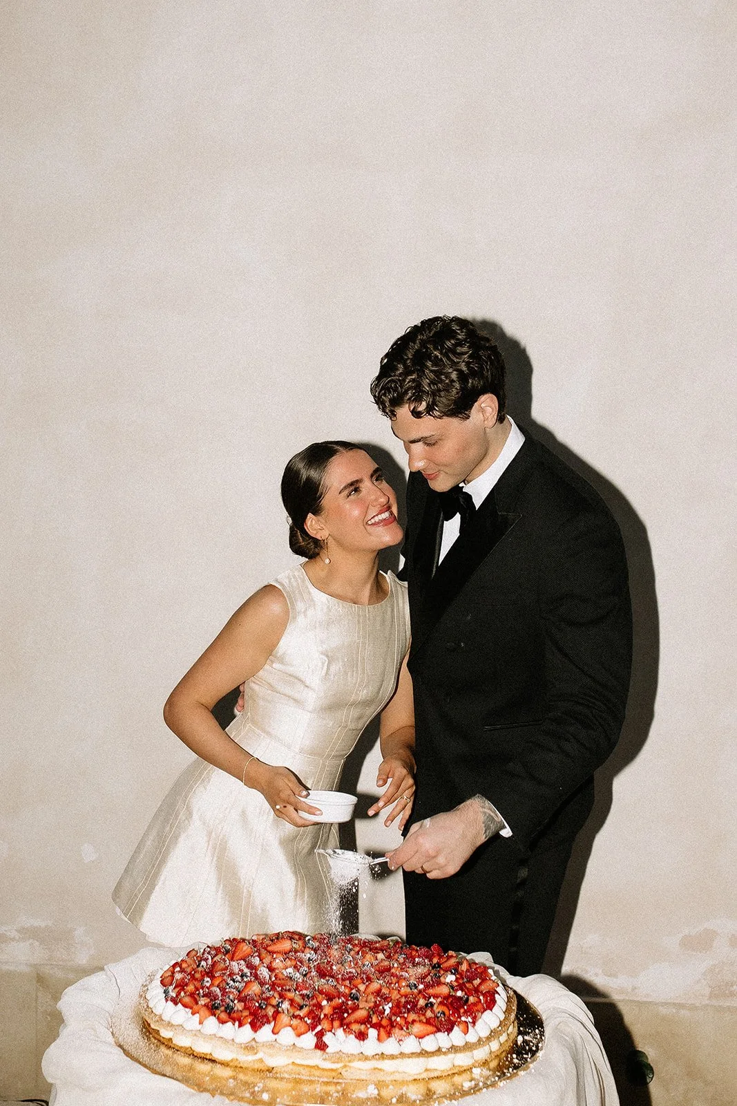 A bride and groom dressed in wedding attire, celebrating with a large strawberry cake, sprinkling powdered sugar, against a plain wall.