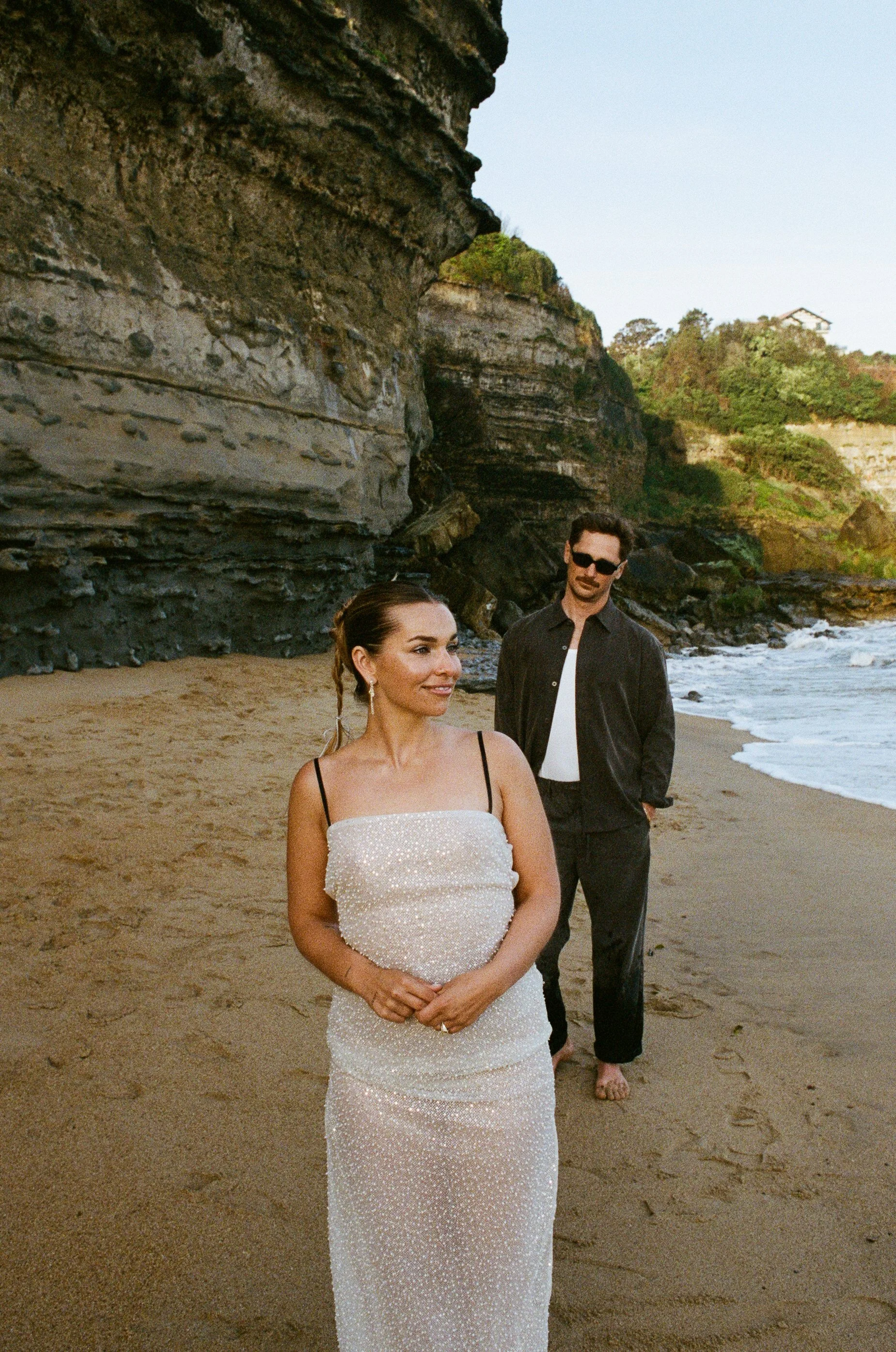 A woman in a white dress with black shoulder straps stands on a sandy beach, smiling, while a man in sunglasses, a dark shirt, and black pants with rolled-up cuffs stands behind her, with a rocky cliff and trees in the background.