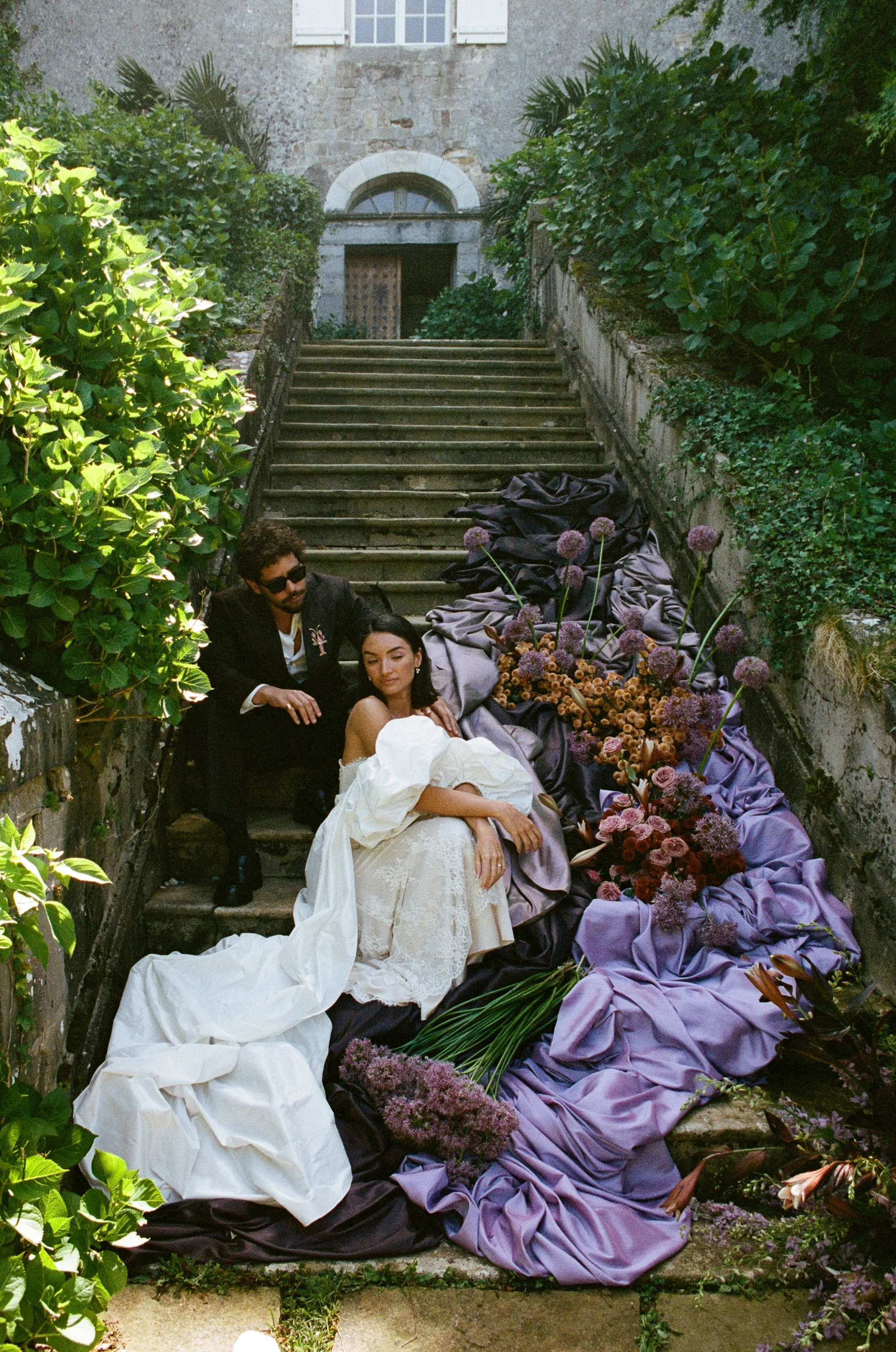 A couple dressed in wedding attire sitting on stone steps surrounded by lush greenery and purple flowers, with a stone building in the background.