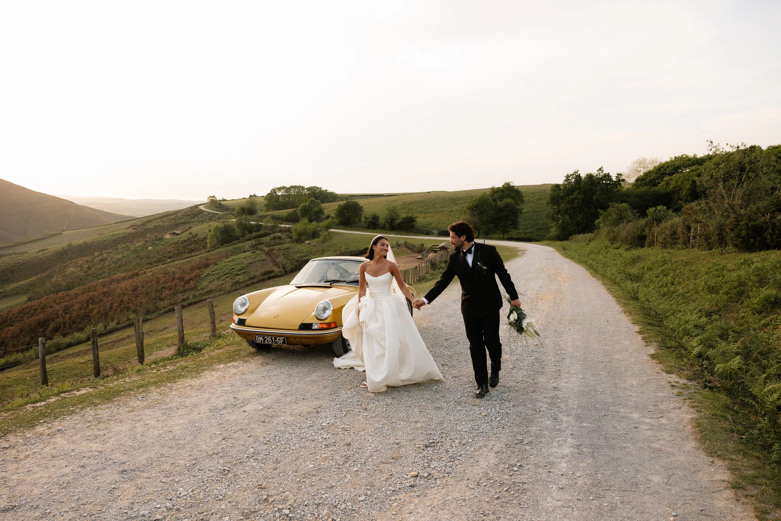 A bride and groom walking hand-in-hand on a gravel road in a rural landscape, with a yellow vintage Porsche parked behind them, rolling hills, trees, and a cloudy sky in the background.