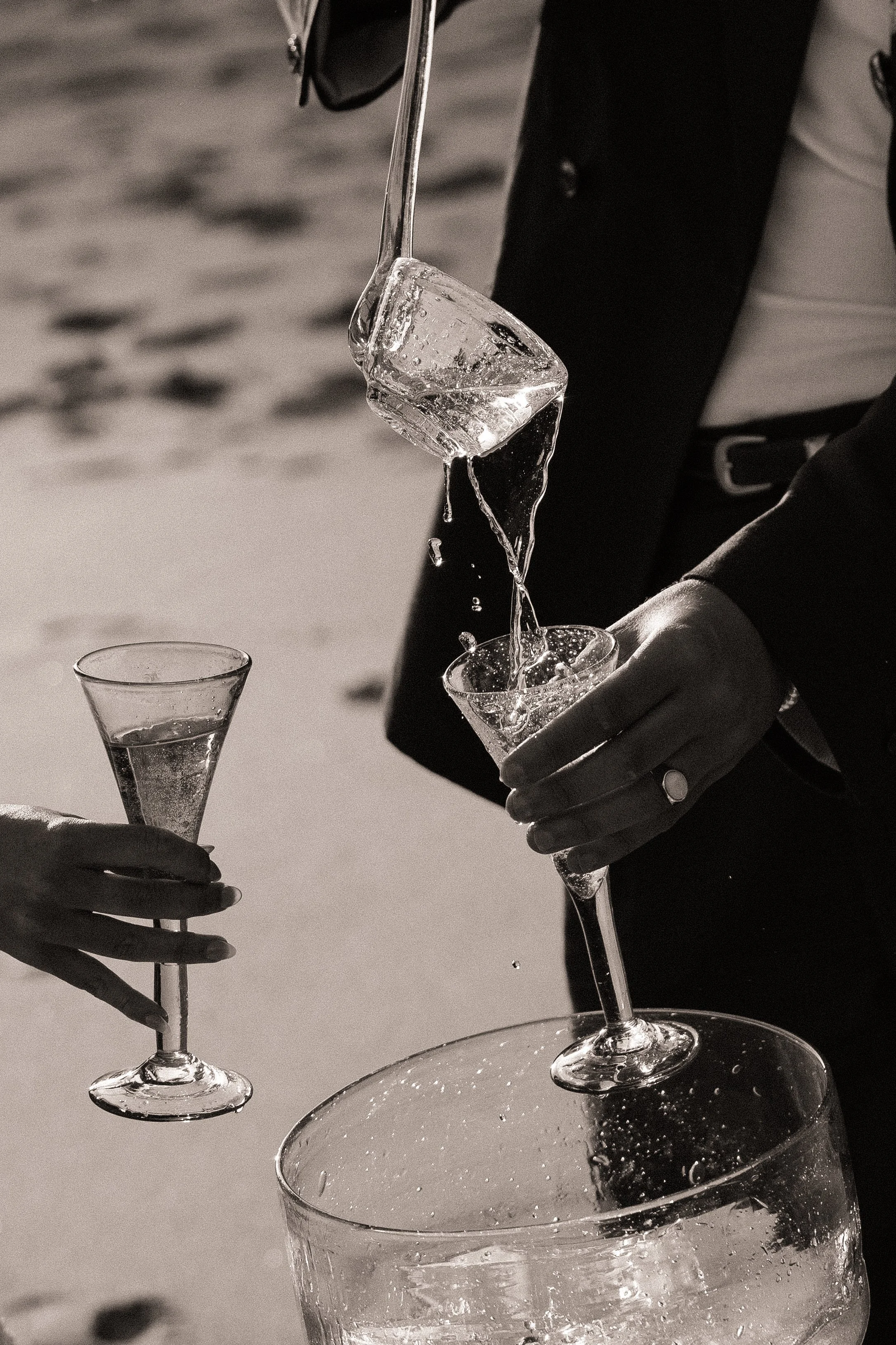Two people are pouring champagne into glasses at a celebration, with one person holding a bottle and the other holding a glass, both wearing formal attire.