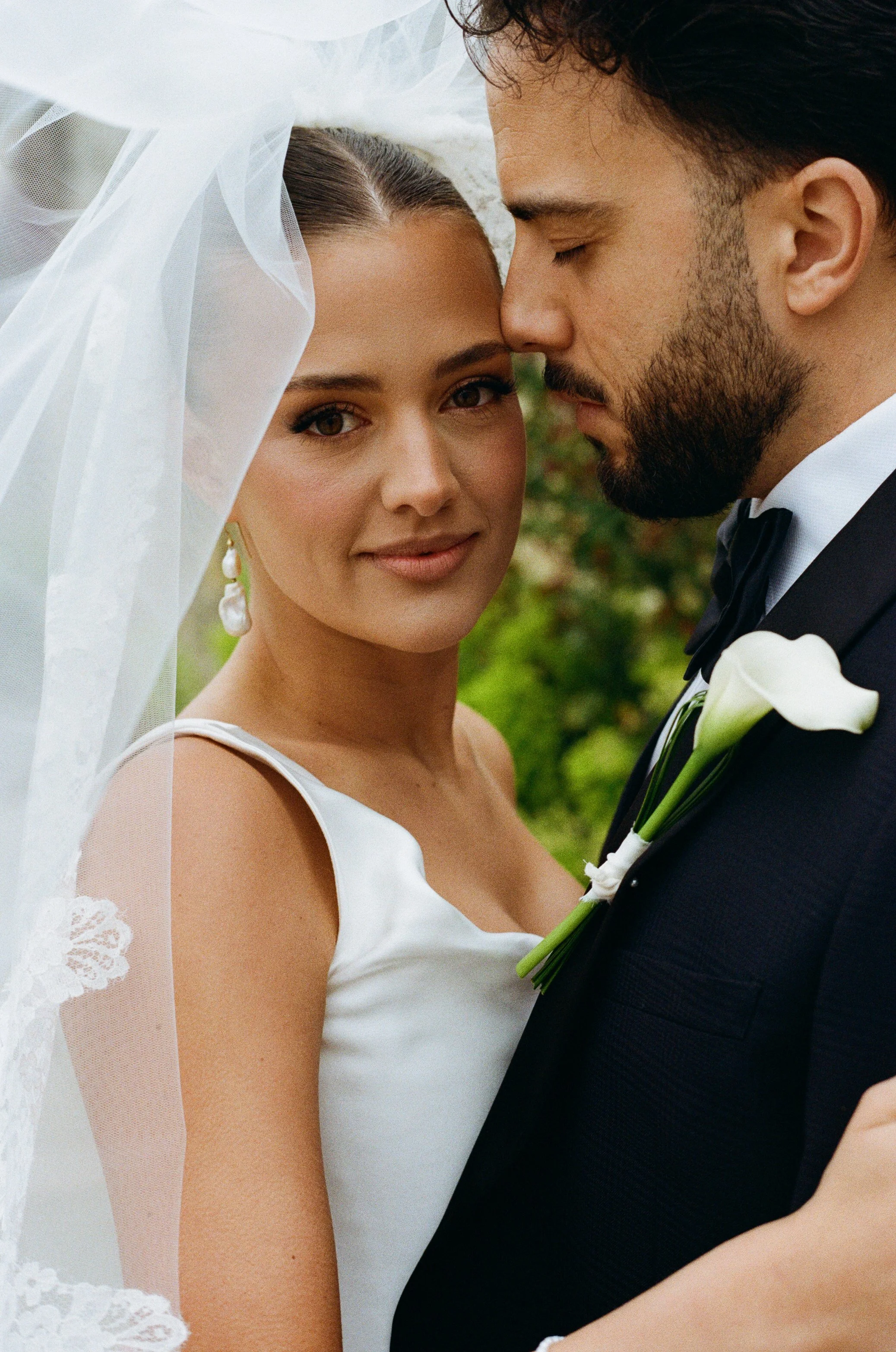 Close-up of a bride and groom with their foreheads touching, dressed in wedding attire, outdoors with green blurred background. Taken on 35mm analogue film.