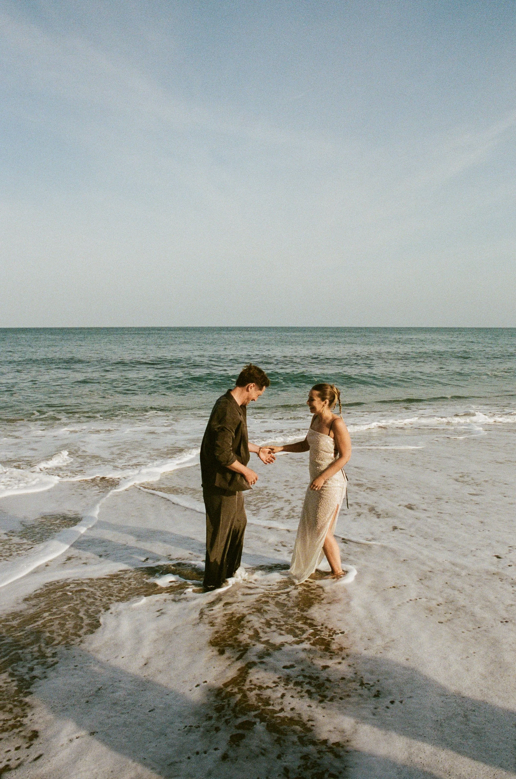 A man and woman are standing on the beach in the ocean, holding hands and looking at each other, with the ocean and sky in the background. Taken on 35mm analogue film.