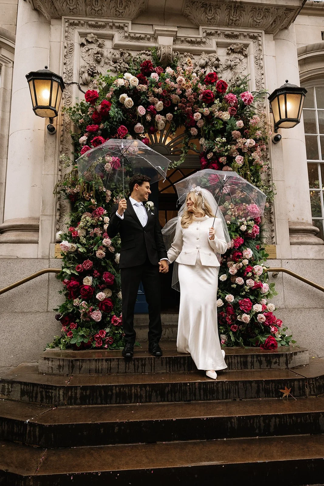 A bride and groom standing on steps outside of Chelsea Town Hall decorated with a large floral arch, holding umbrellas, smiling at each other.