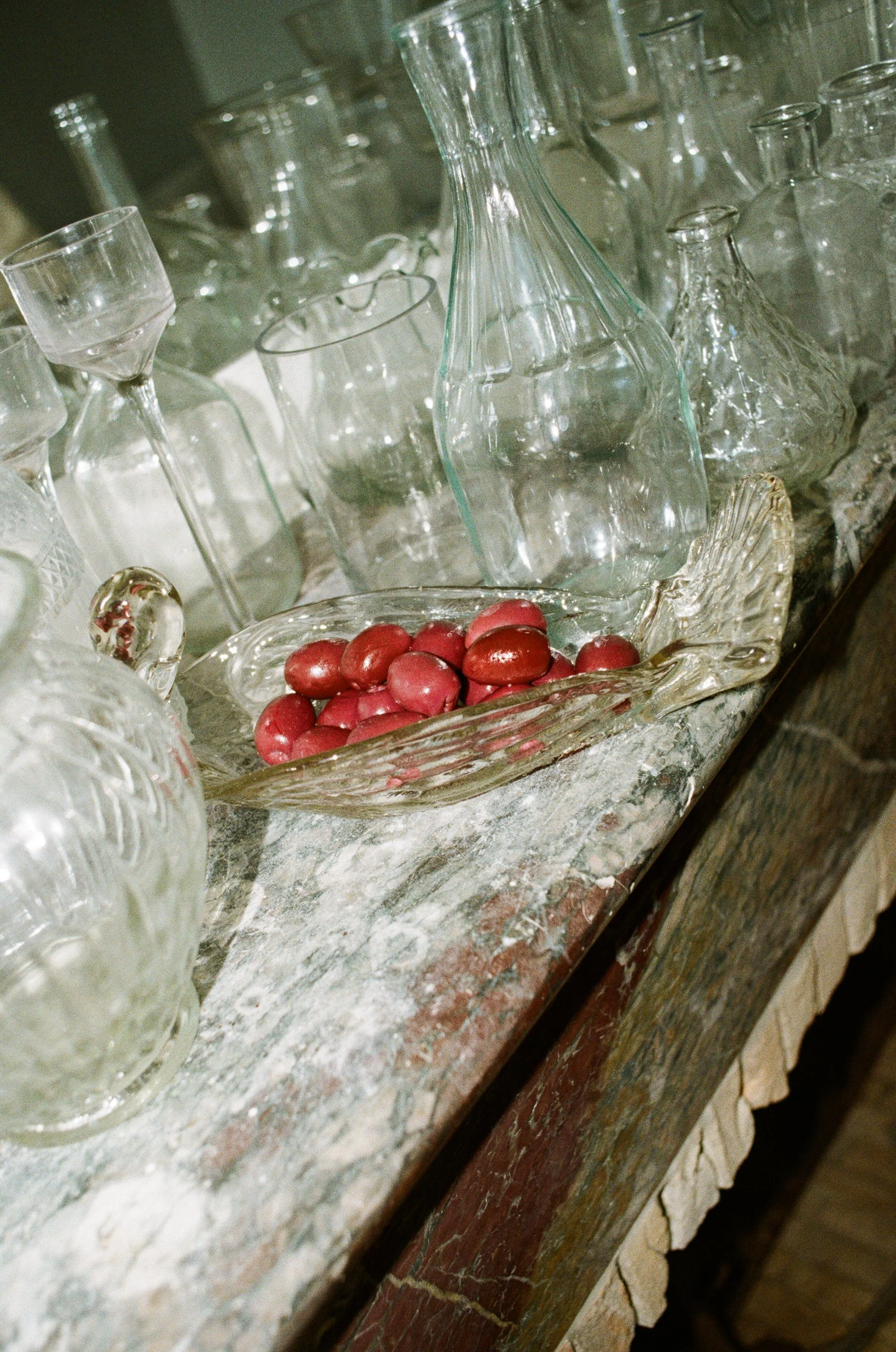A display of various glass decanters and bottles on a marble countertop, along with a bowl of red grapes.