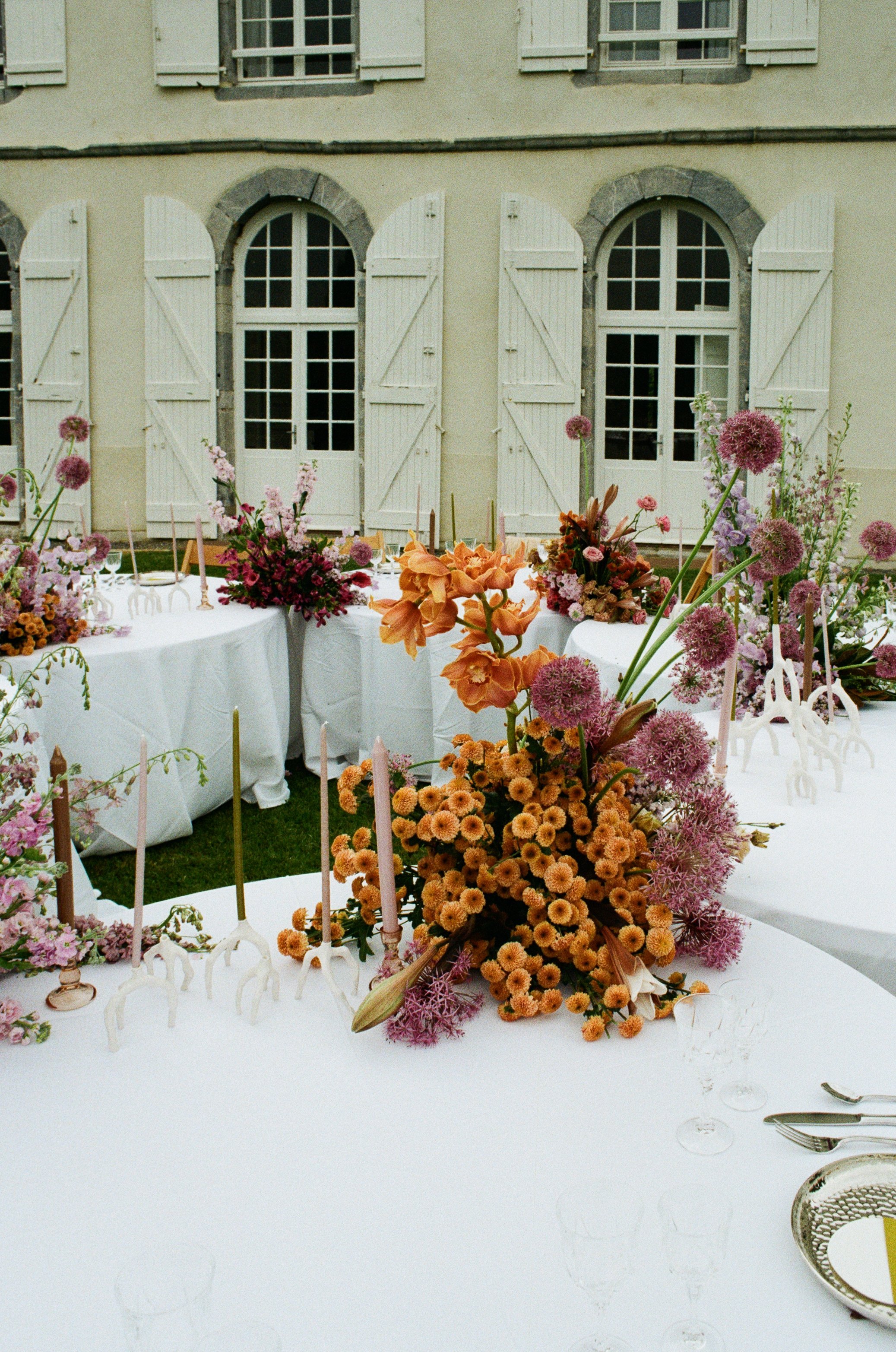 Elegant outdoor dining setup with white tables and floral centerpieces featuring pink, orange, and purple flowers, set against a yellow building with arched windows and white shutters.