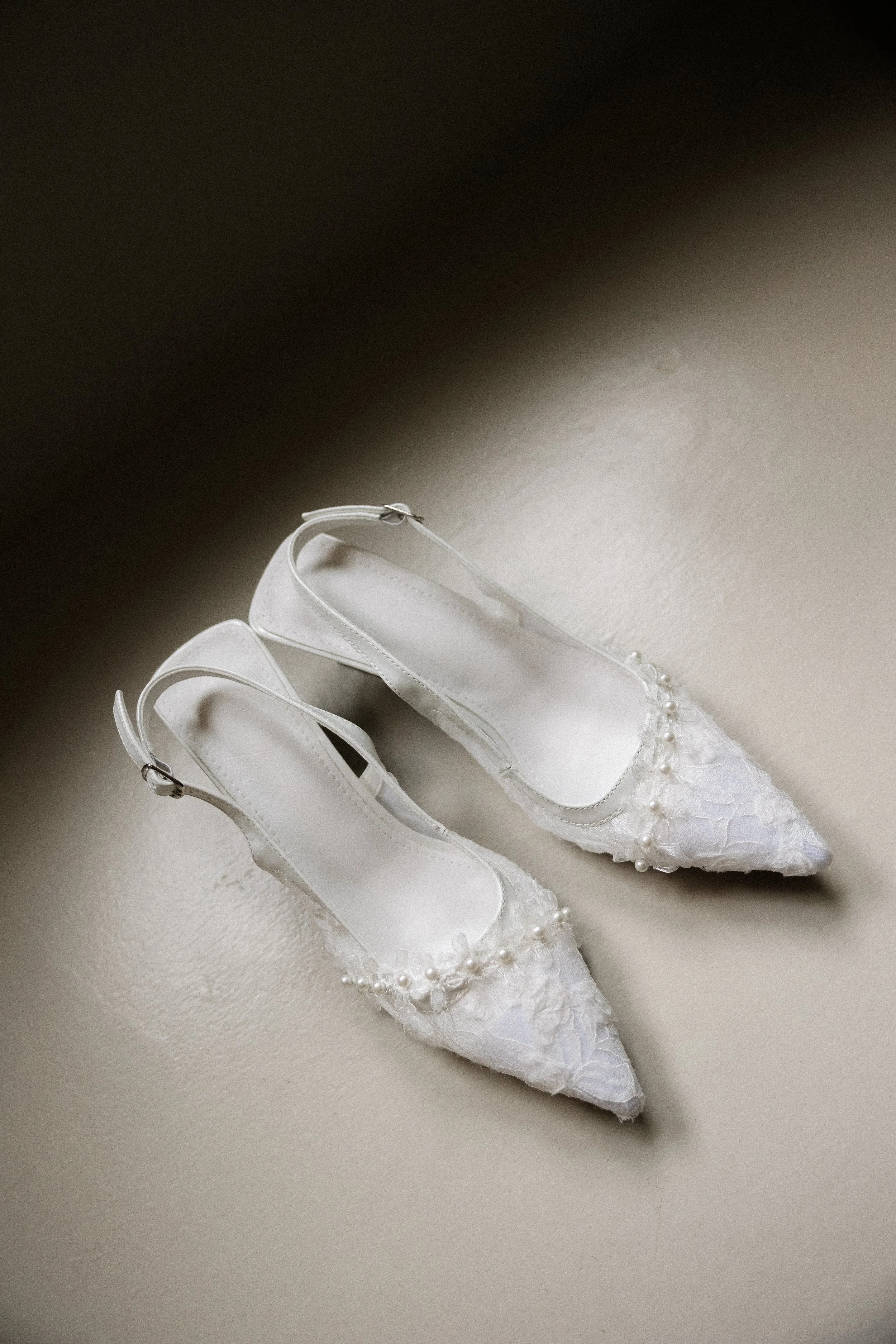 A pair of white bridal shoes with ankle straps, decorated with pearls and lace, placed on a light-colored surface.