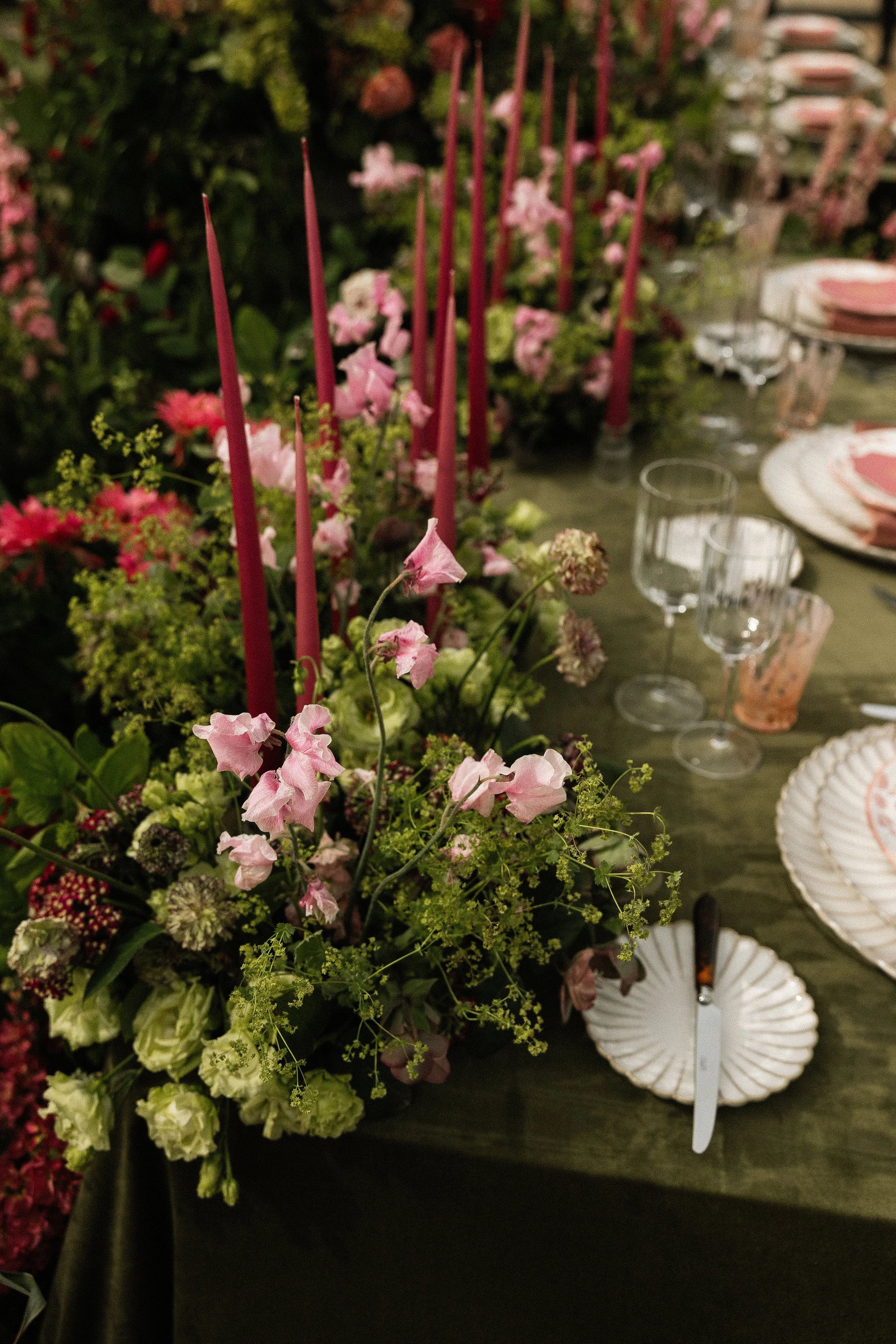 A floral centerpiece with pink and white flowers and tall pink candles on a dining table, with place settings and glassware visible in the background.