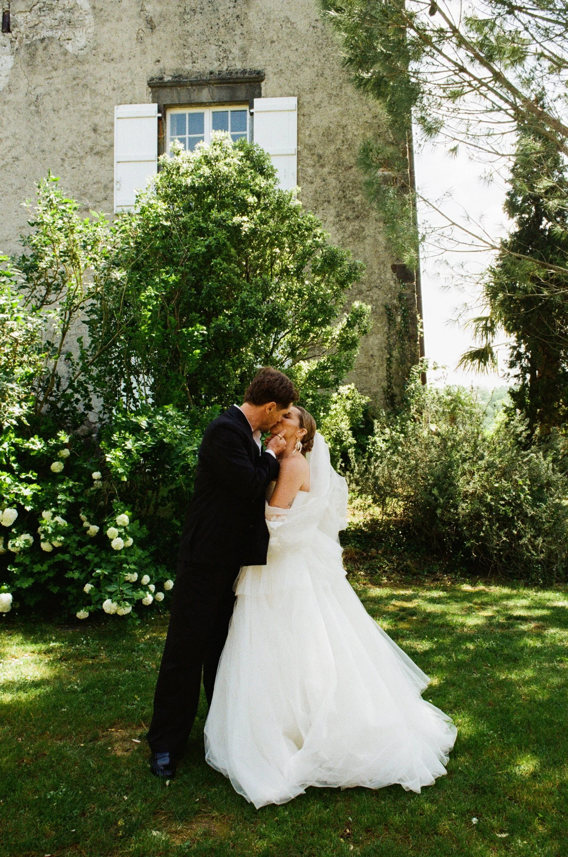 A bride and groom sharing a kiss outdoors in a garden, with green foliage and a stone building with white shutters in the background.
