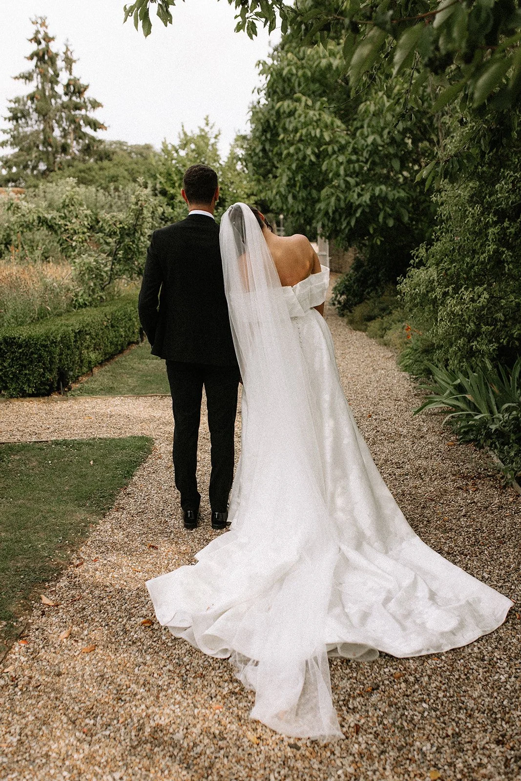 A bride in a white wedding gown and veil walking with a groom in a black suit along a garden pathway.