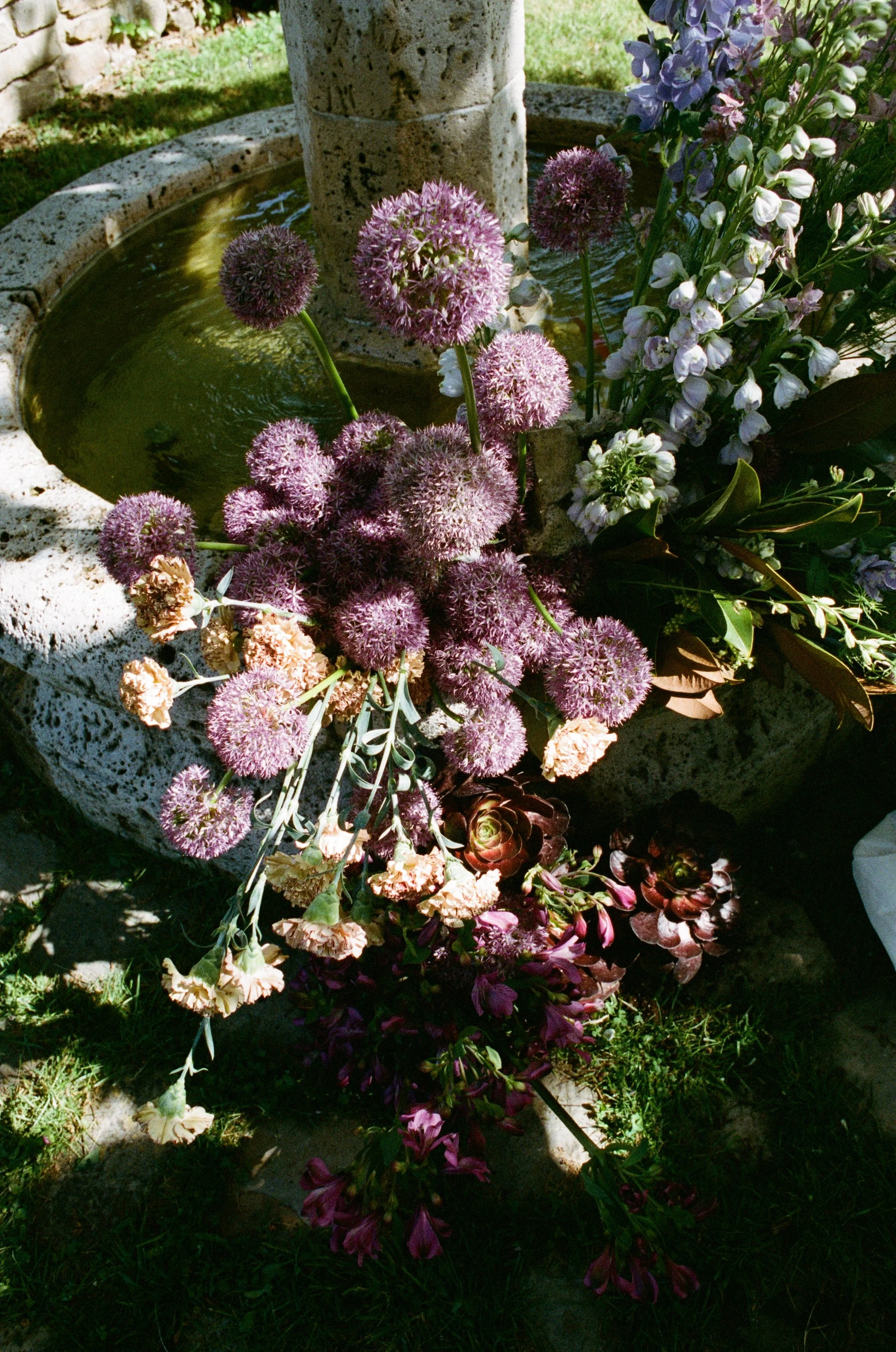 A circular stone flower fountain with a small water basin, surrounded by purple, white, and pink flowers and green grass.