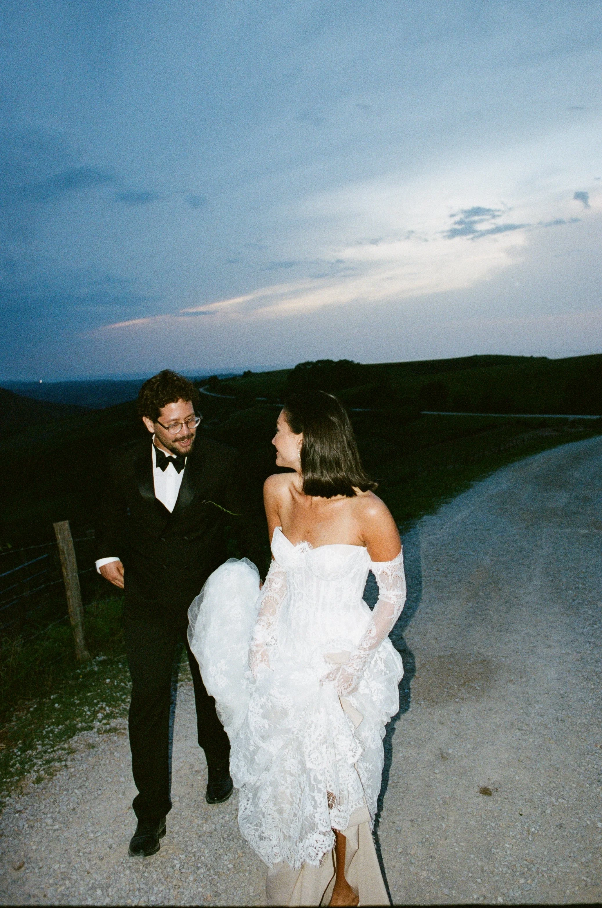 A man in a tuxedo with glasses and a woman in a strapless lace wedding dress with an off-the-shoulder neckline, standing outdoors on a gravel path near a river with rolling hills in the background during dusk. Taken on 35mm analogue film.