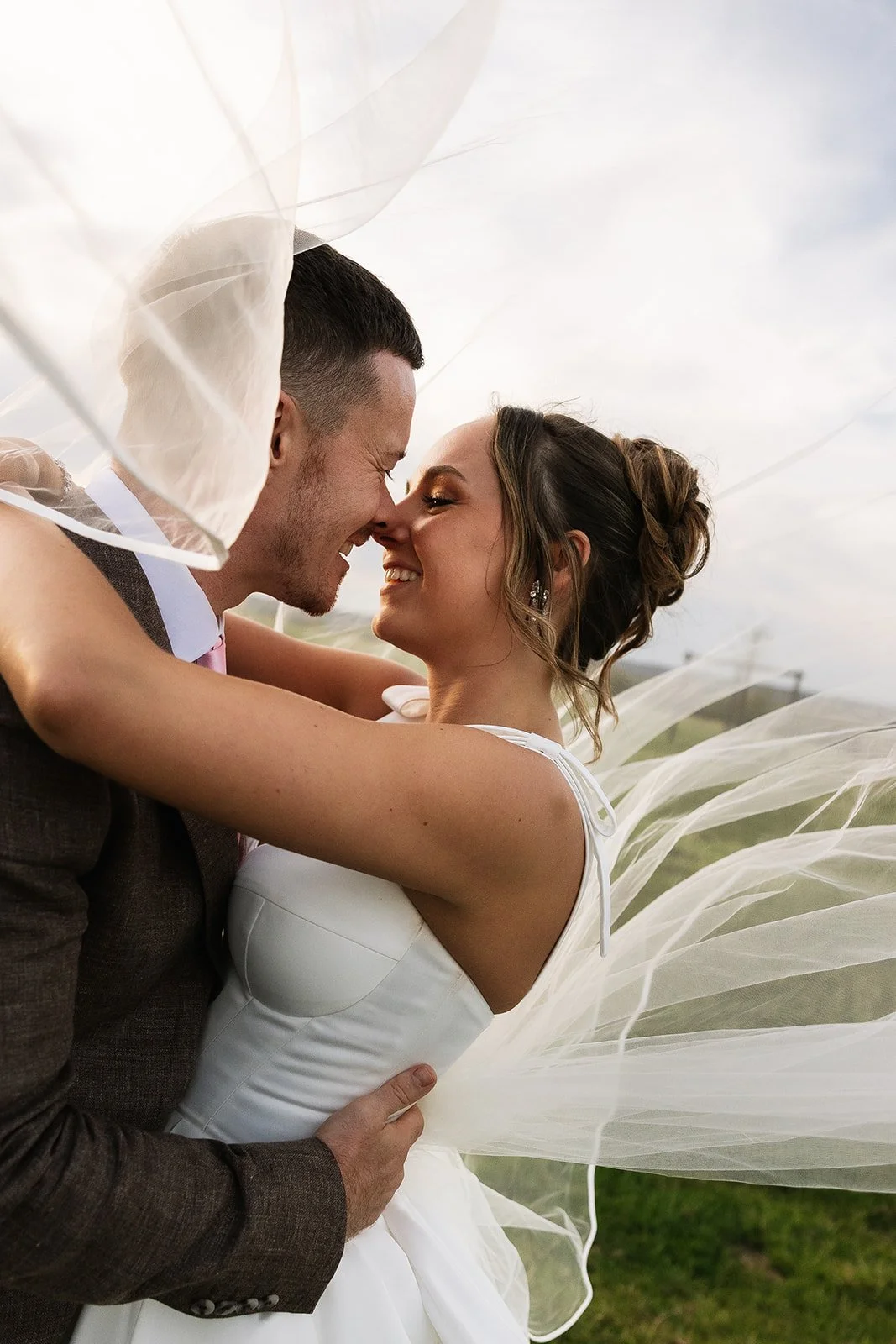 A bride and groom embrace outdoors, touching noses and smiling, with the bride's veil flowing around them.