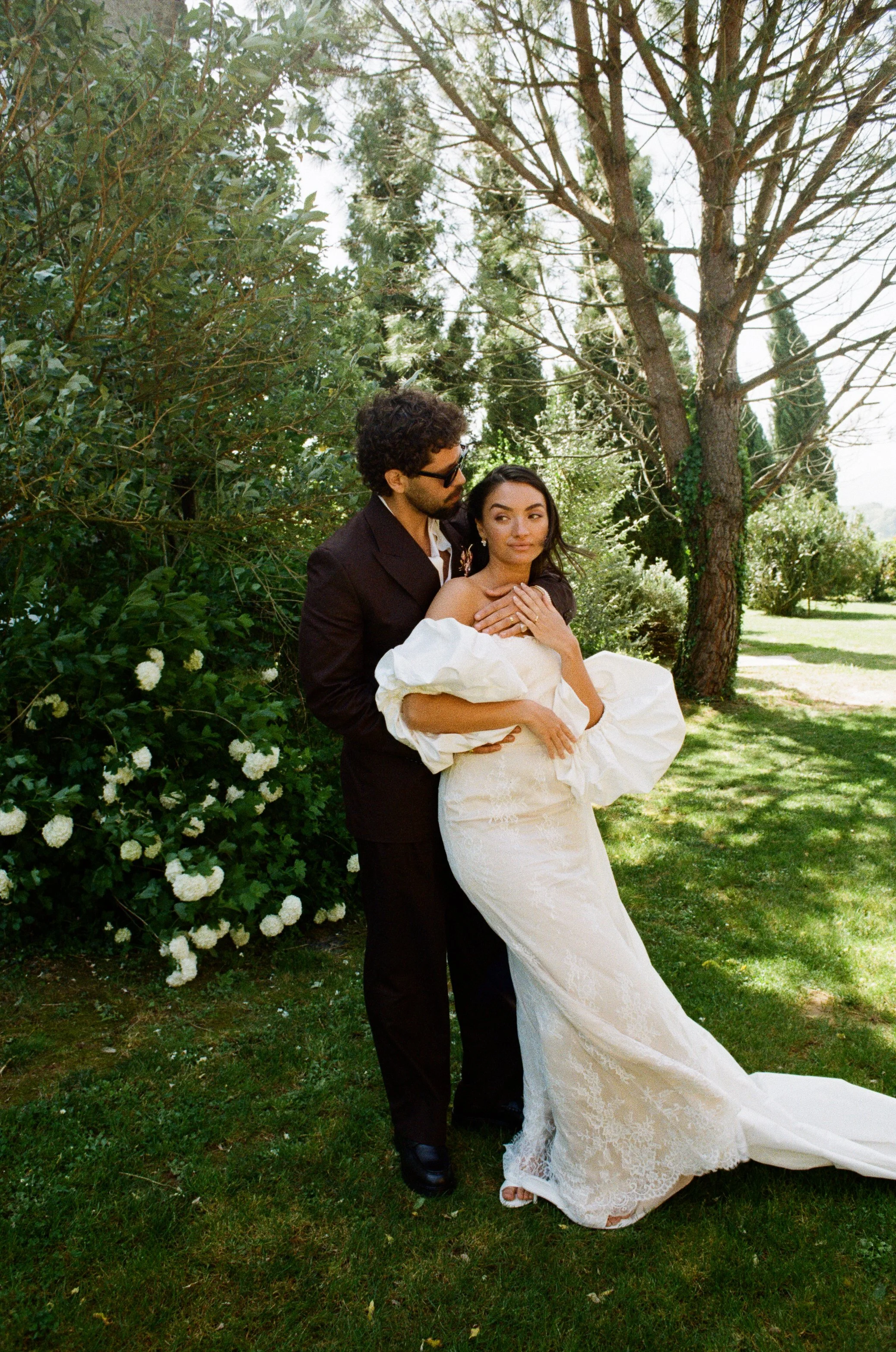 A couple, dressed in a wedding gown and tuxedo, pose outdoors in a garden with trees and greenery.