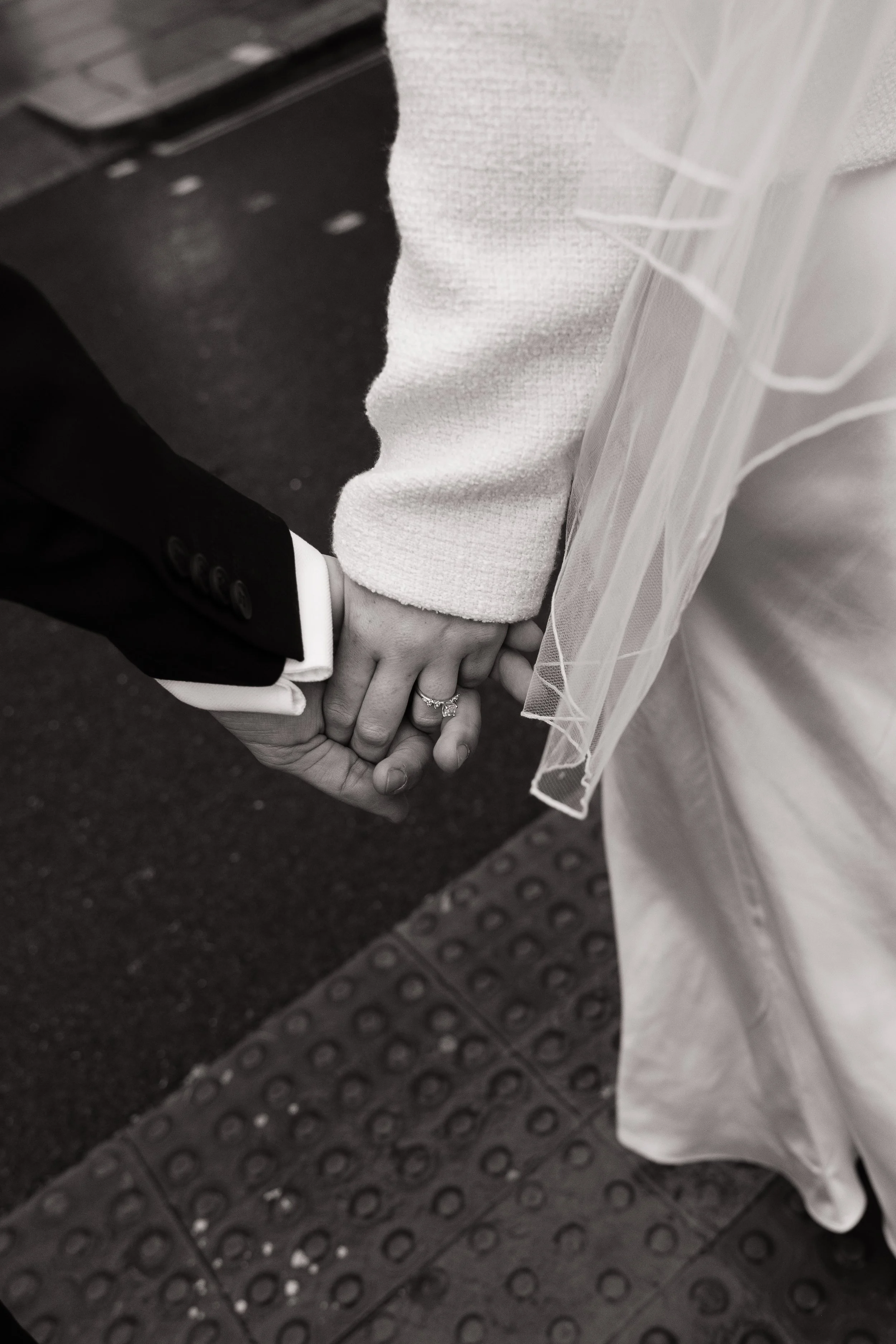 A close-up photo of a bride and groom holding hands, with the bride wearing a white wedding dress and veil, and the groom dressed in a black suit with white shirt and buttons visible on the sleeve, standing on a rubber mat.