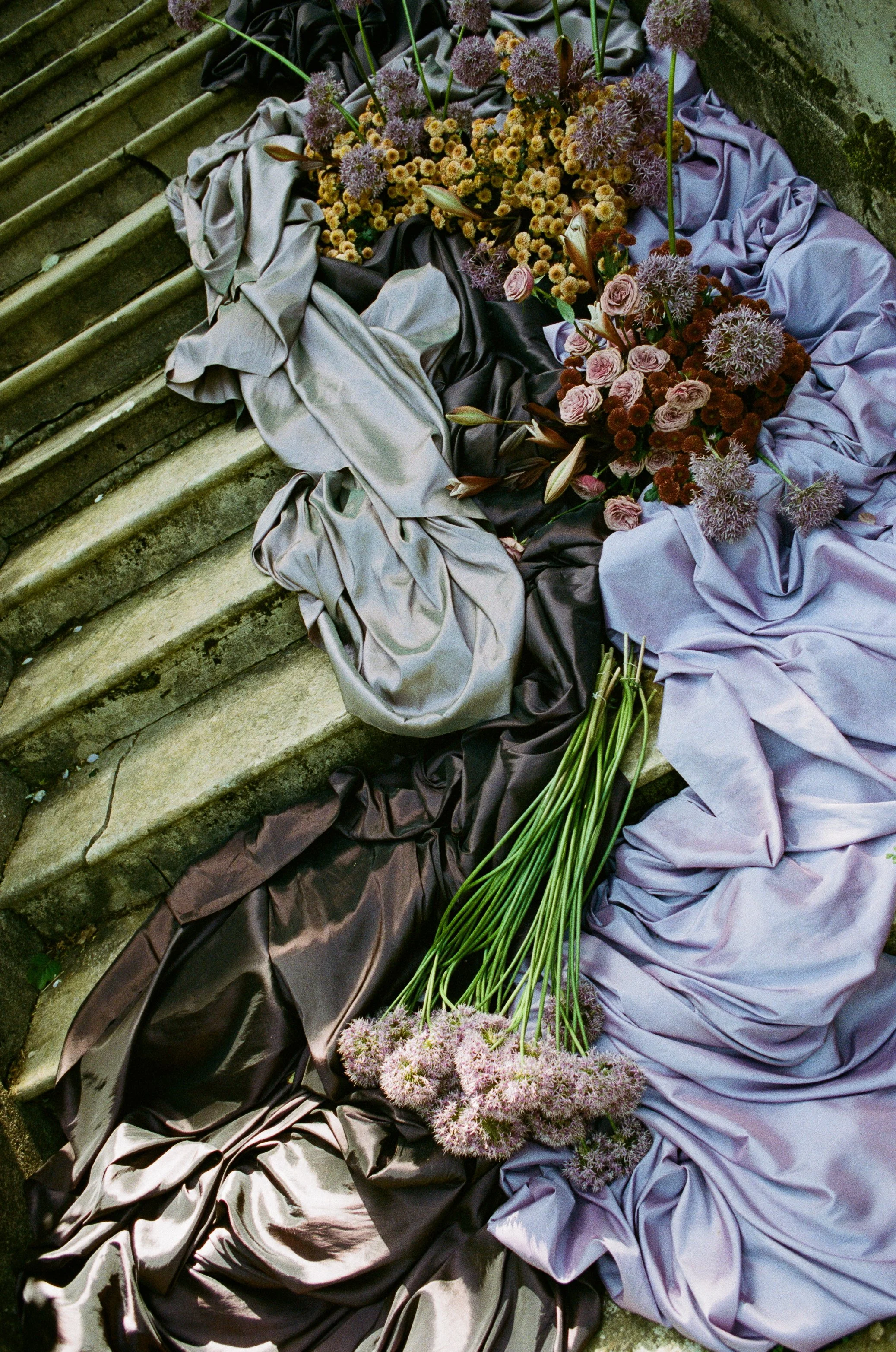 An arrangement of flowers on a stone staircase, with silk fabric in shades of silver, black, and lavender underneath. Taken on 35mm analogue film.