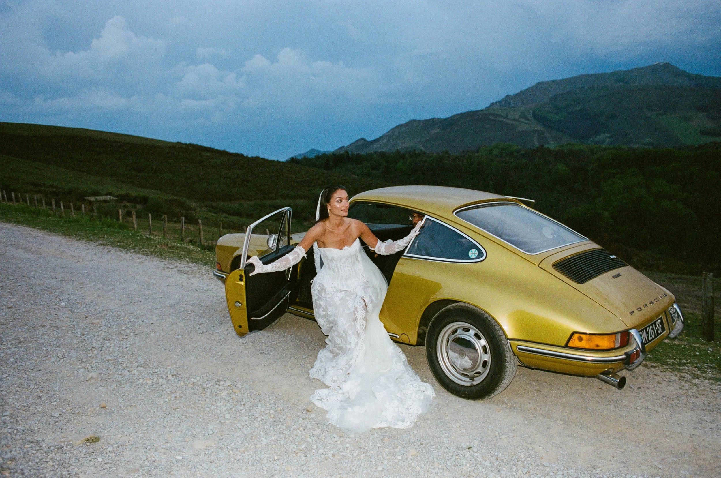 A woman in a white wedding dress is standing by a yellow Porsche car with the door open in a rural landscape with green hills and mountains in the background during daytime.